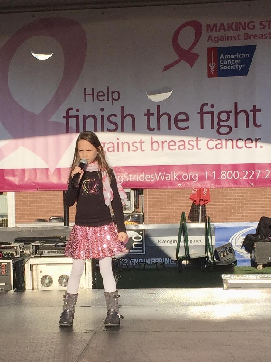 Young girl standing in front of a breast cancer awareness banner, wearing a pink skirt, black top, and boots.