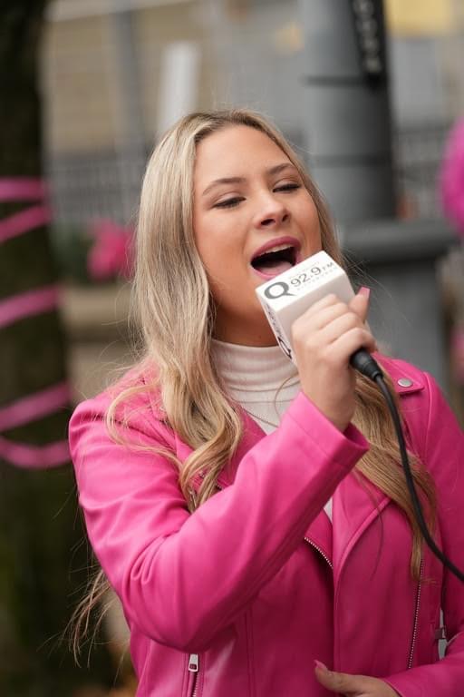Young woman with long blonde hair singing into a microphone outdoors, wearing a pink jacket and white turtleneck, with blurred background.