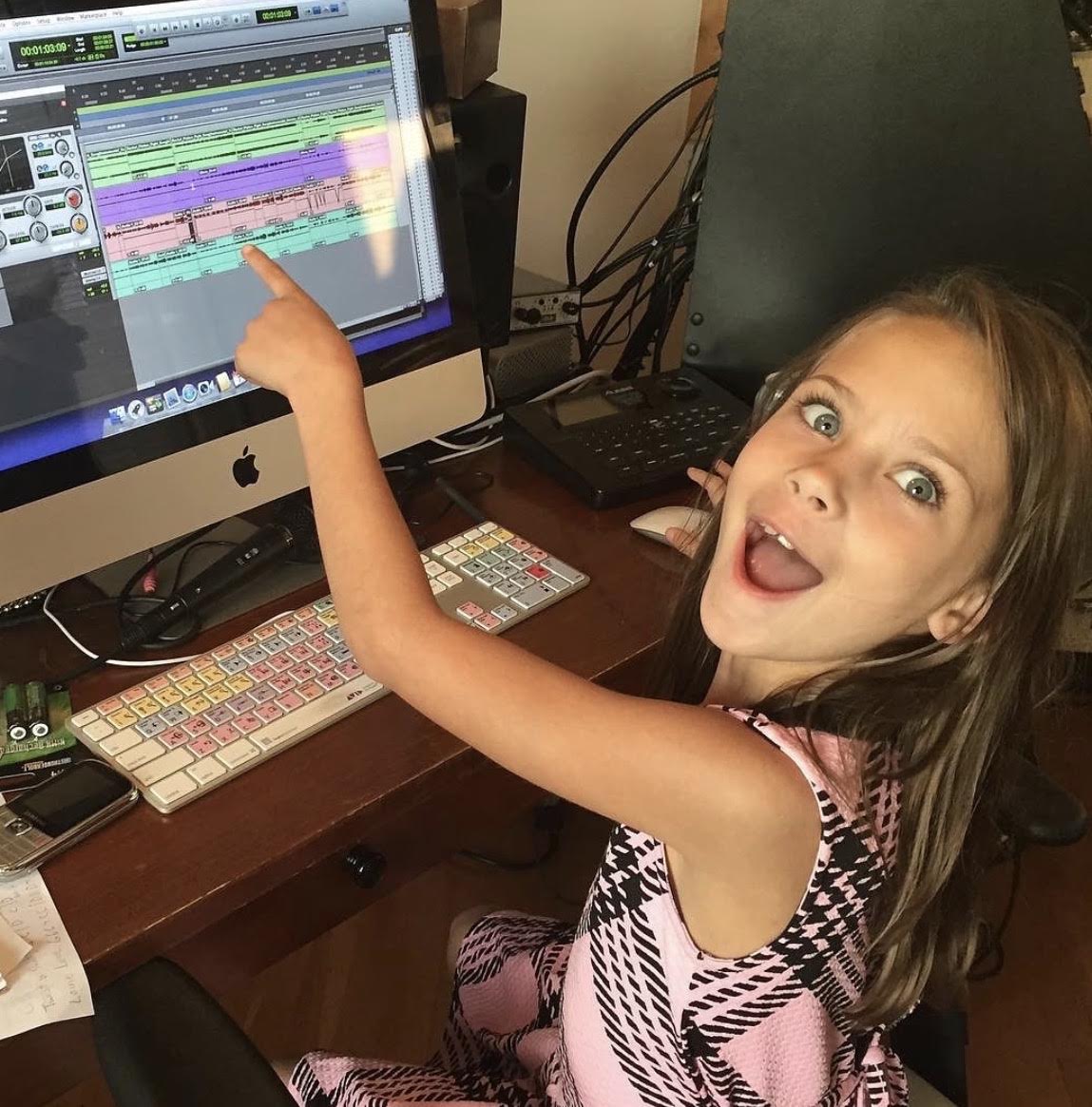 Young girl with long hair pointing at a computer screen showing a music editing program, smiling and sticking out her tongue.