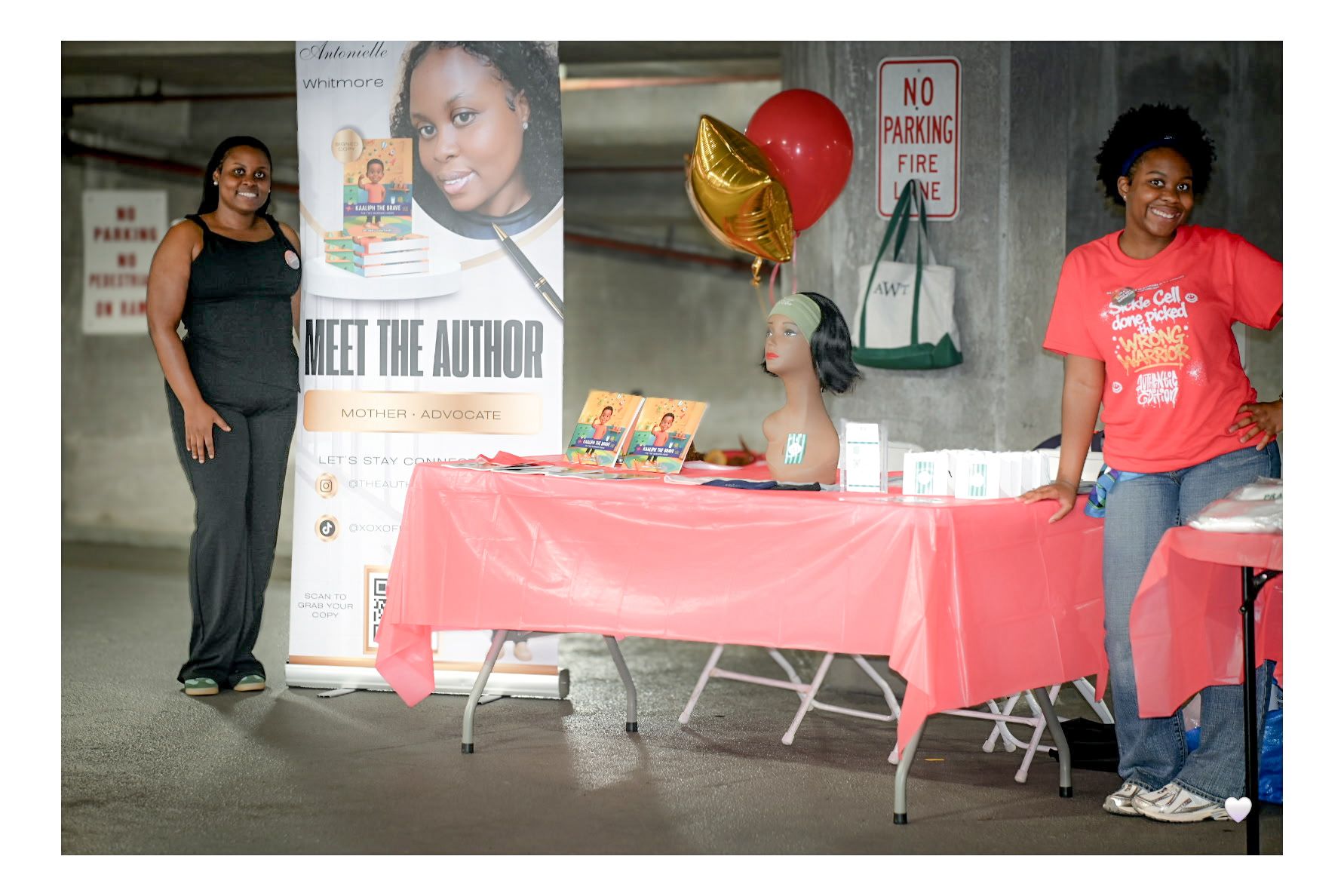 Two women stand near a table with balloons and books, one sitting behind the table, in an indoor parking area.