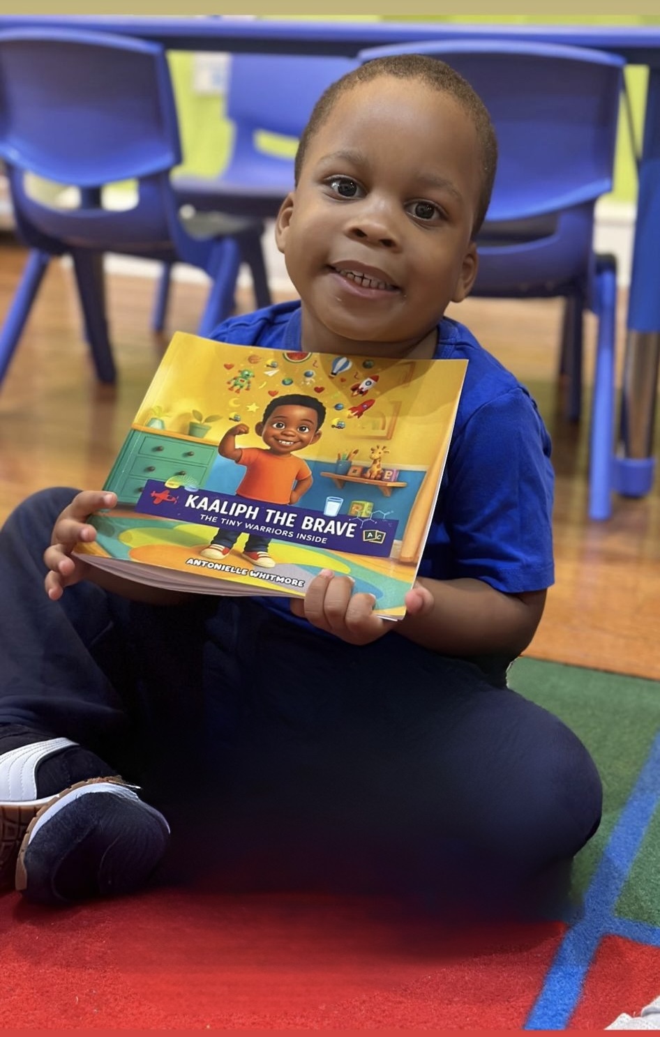 Young child sitting on the floor holding a colorful book, smiling, with chairs in the background.