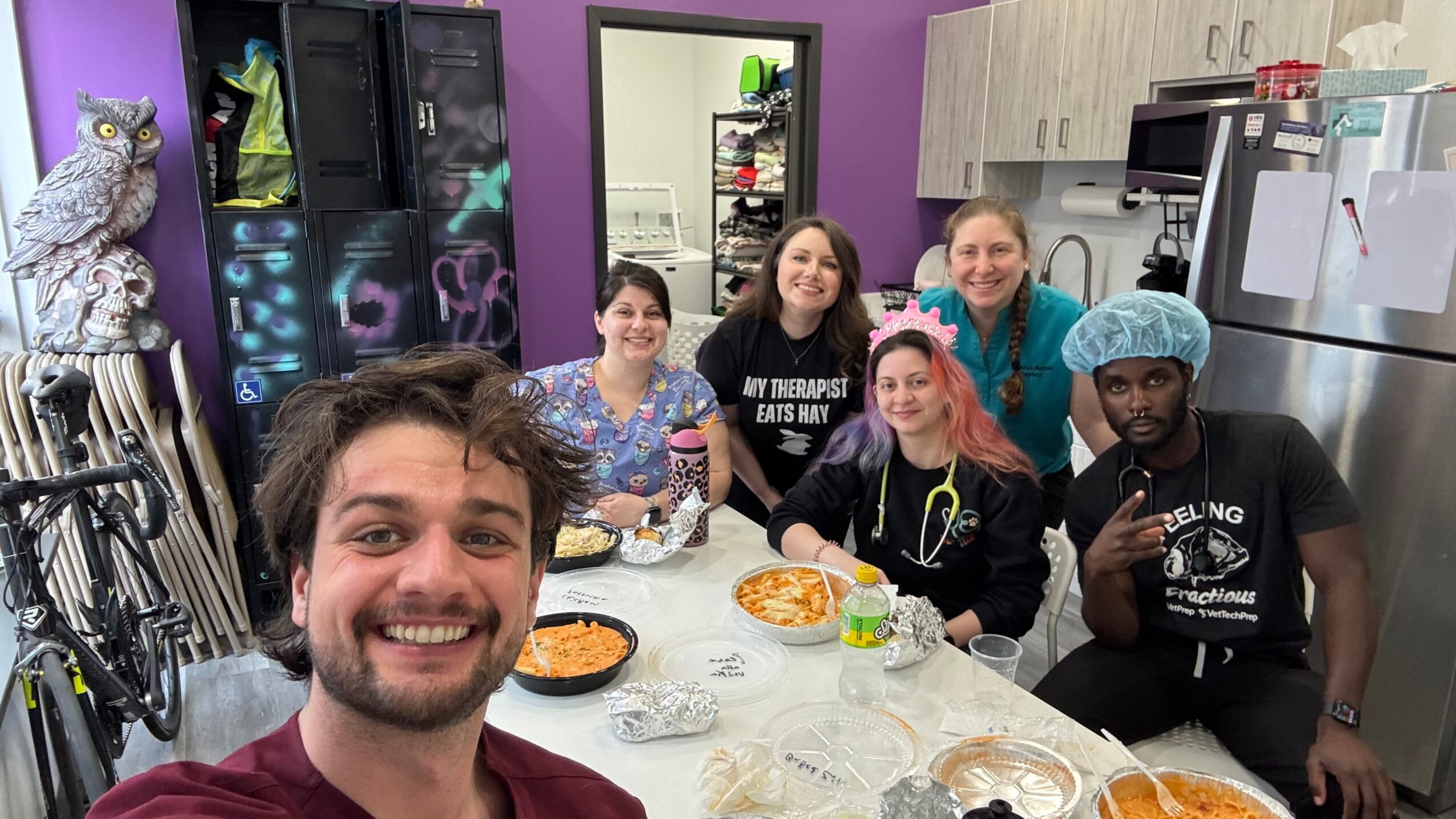 Group of six people in a kitchen, smiling around a table with snacks, with a person taking a selfie. Colorful background.