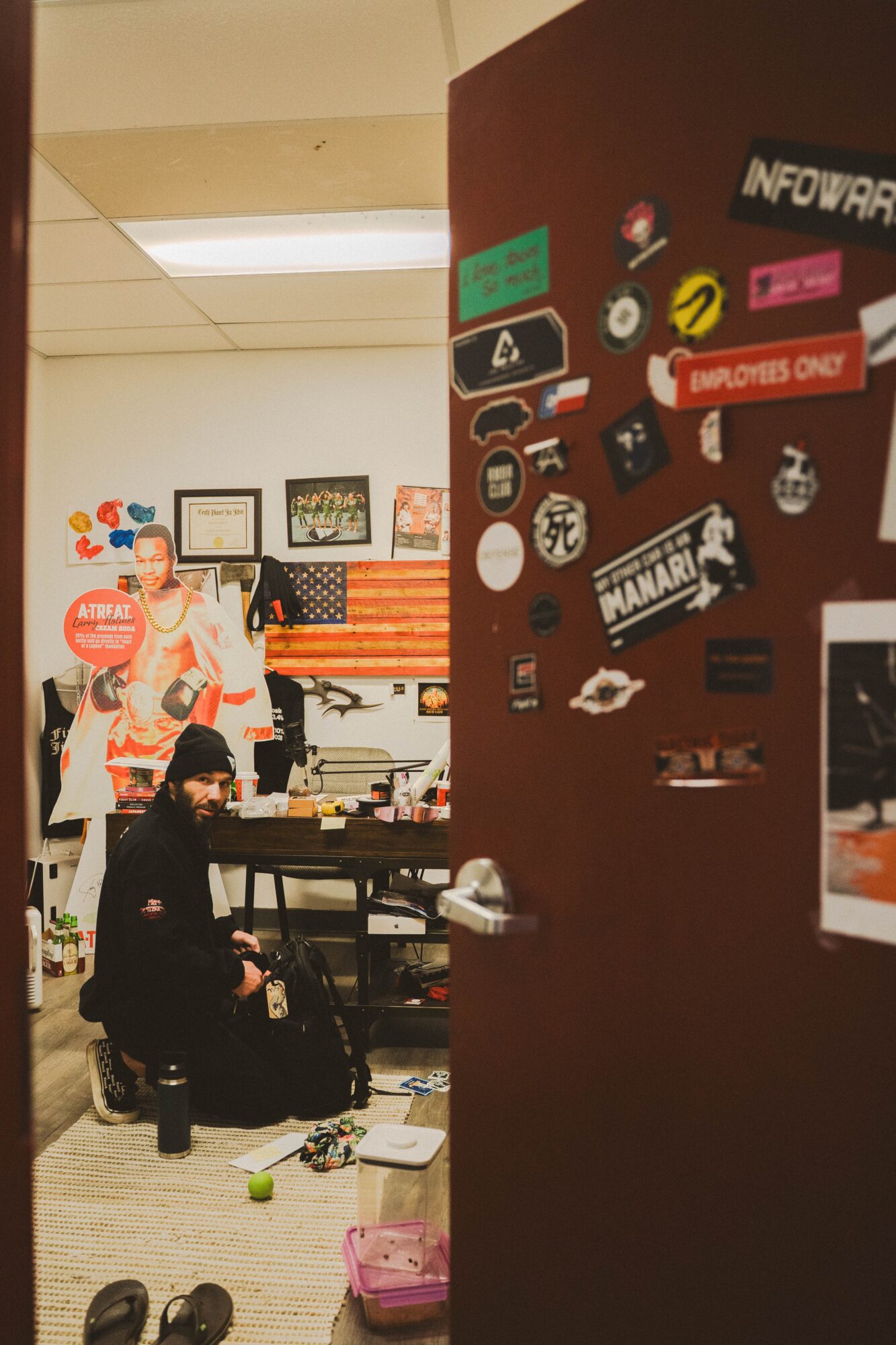 Person sitting at a desk in a cluttered room with a wall covered in stickers and signs, and another person in the background.