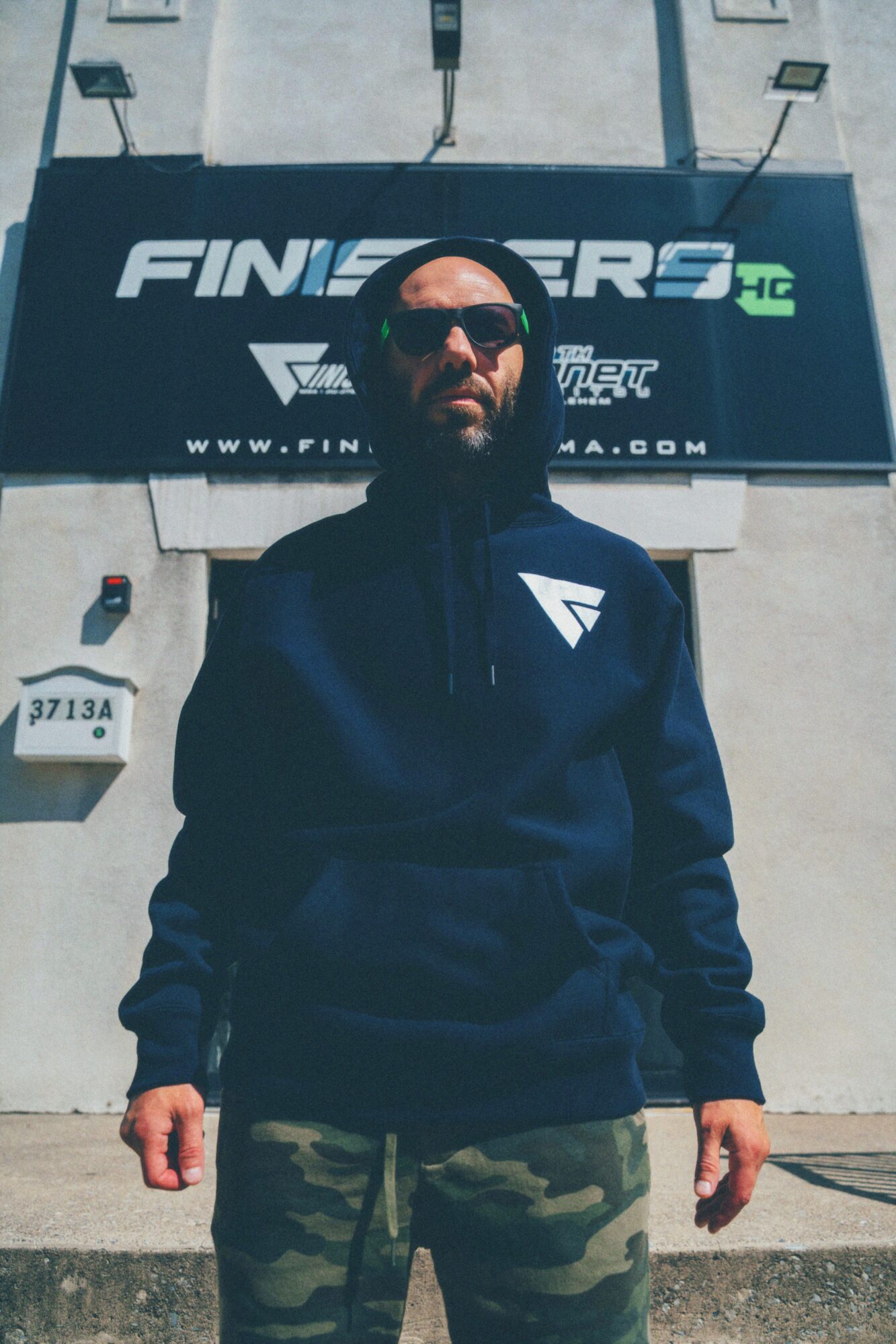 Man wearing sunglasses and a black hoodie with a logo, standing in front of a sign with text, outdoors.