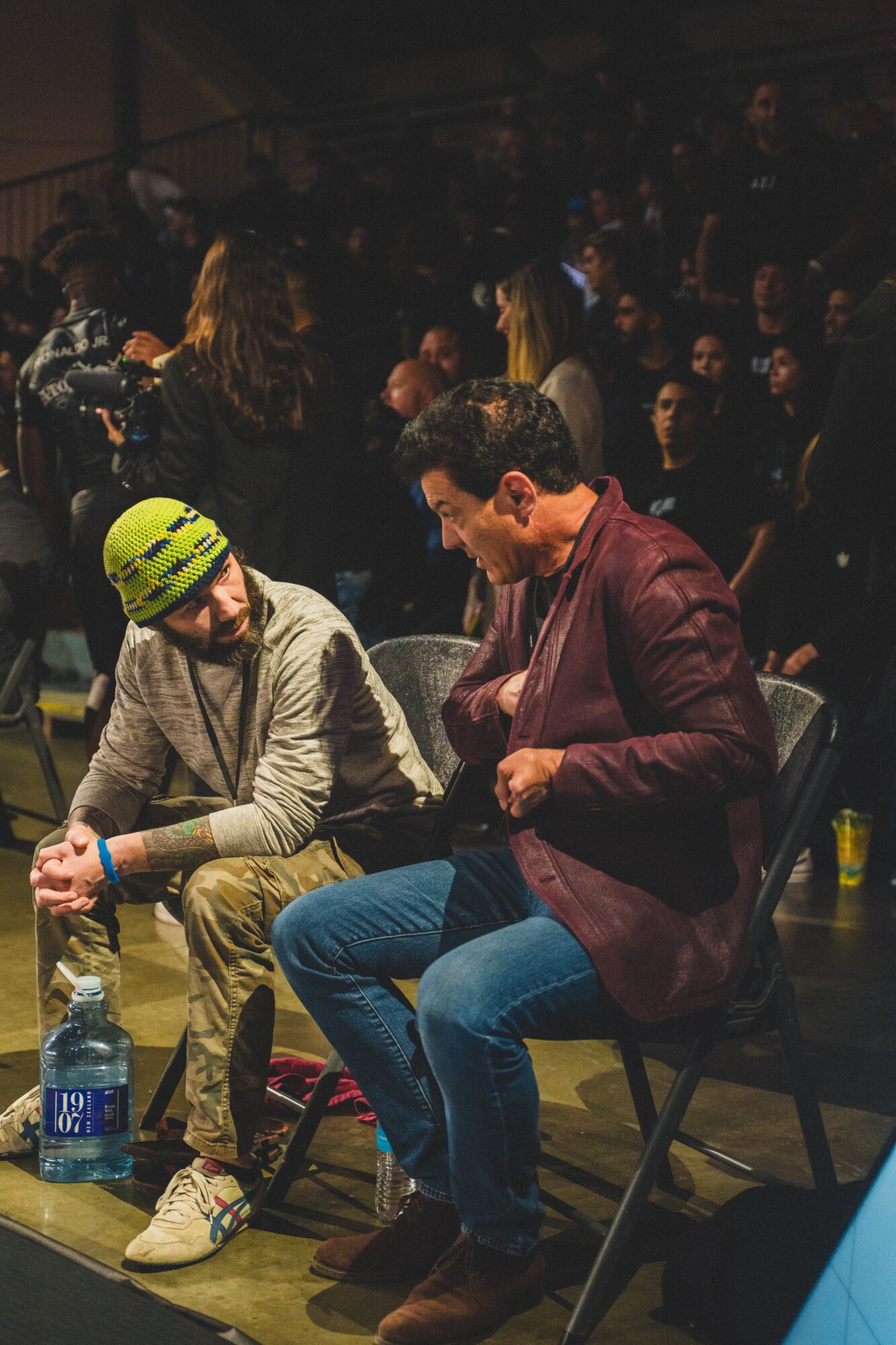 Two men sit on chairs in a crowded indoor setting, one wearing a yellow helmet and the other in a maroon jacket, with people in the background.