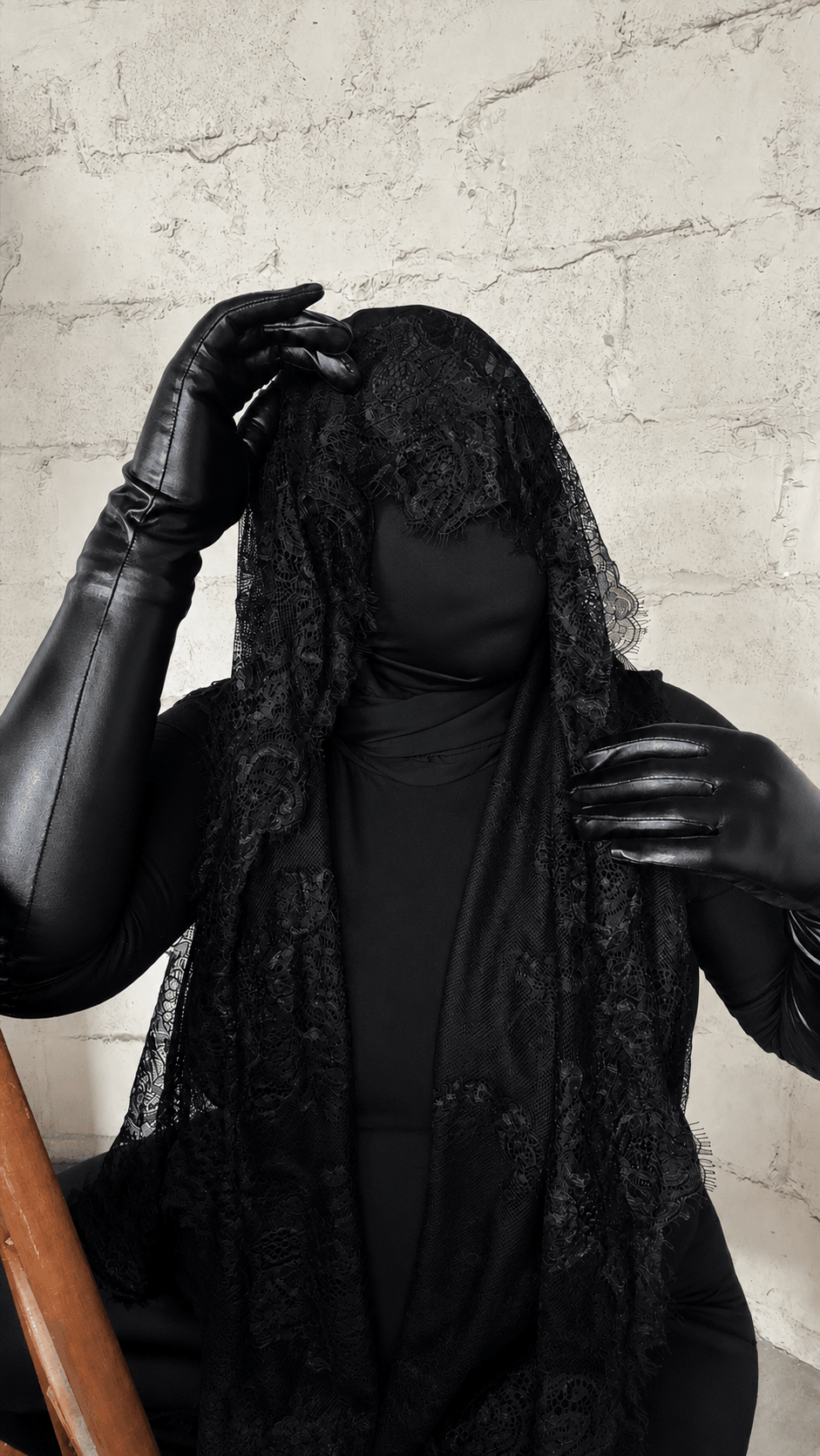 Person with curly hair wearing black clothing and gloves, standing against a textured wall.