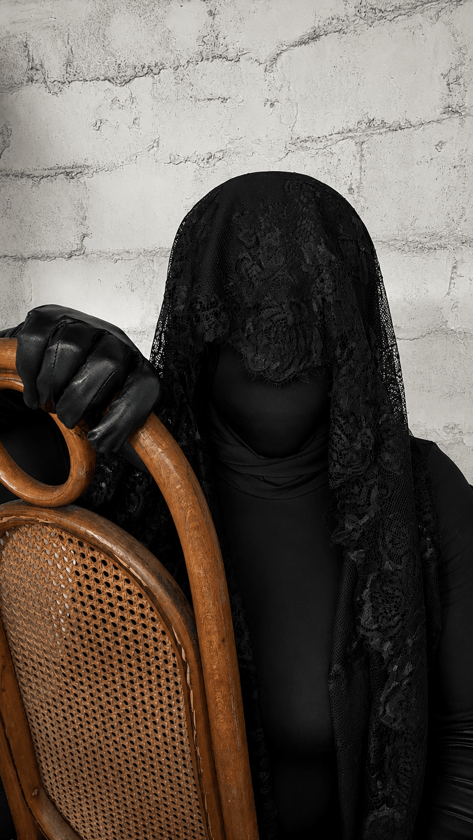 Person with face covered by black lace veil, holding a cane, against a white brick wall.