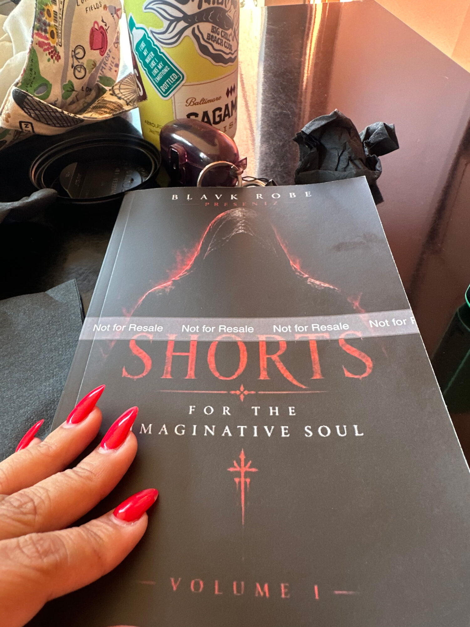 Person with red painted nails holding a book titled 'Shards for the Imaginative Soul, Volume 1'. Background includes cans and a black object.