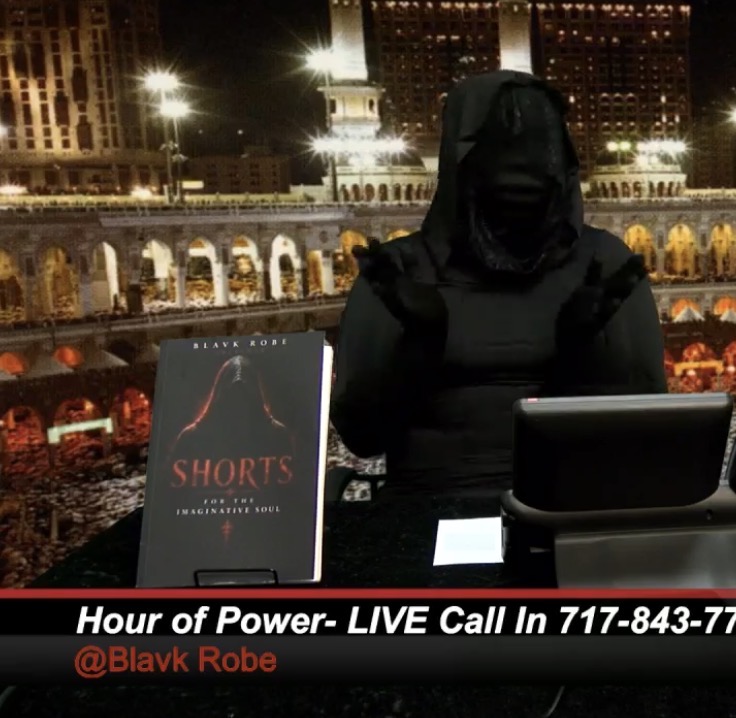 Person in black hoodie sitting at a table with a laptop and a book titled 'Shorts' outdoors at night, city lights in background.