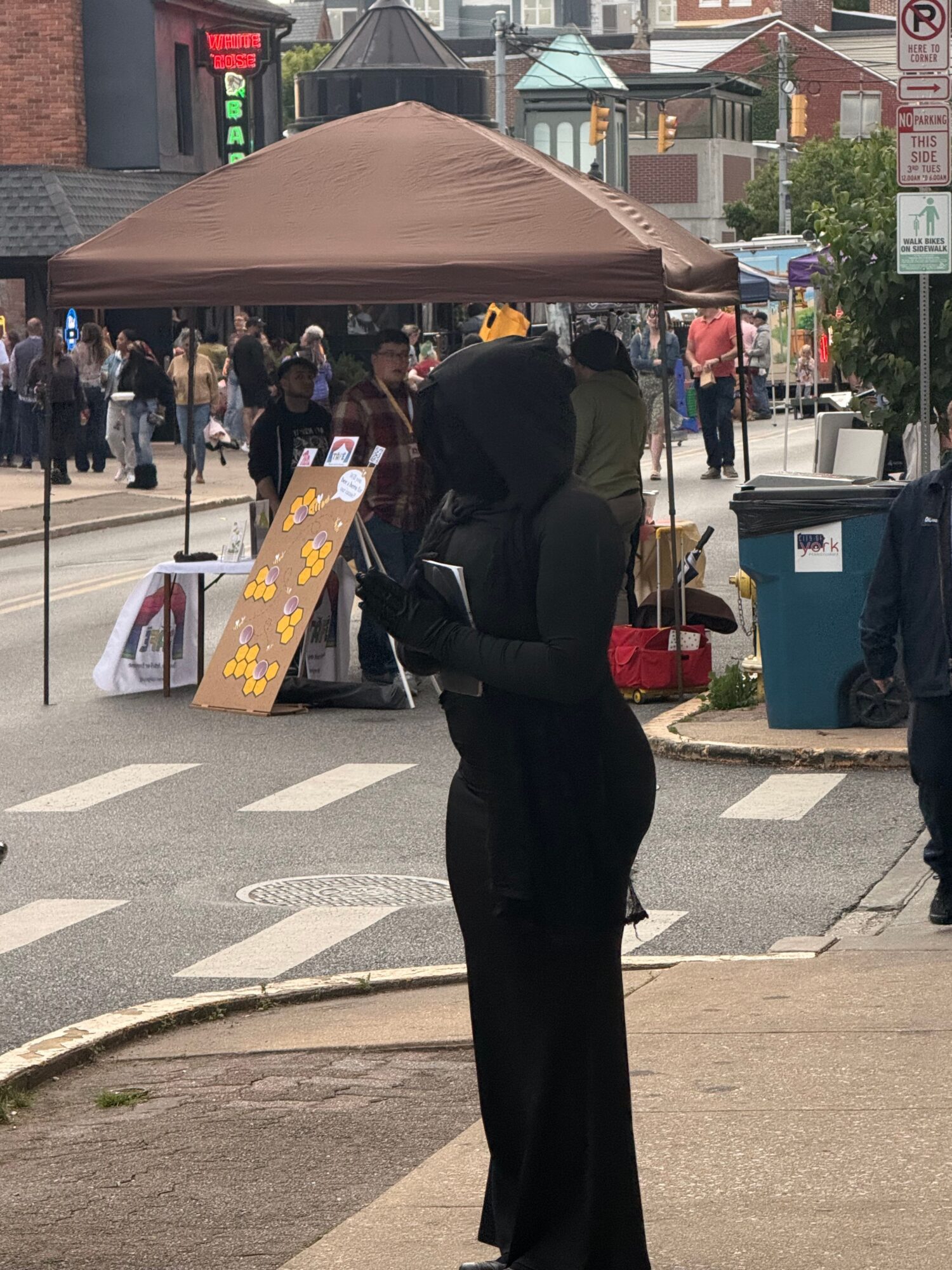 Person standing under a canopy on a busy street with shops and pedestrians, holding a bag, with a signboard nearby.