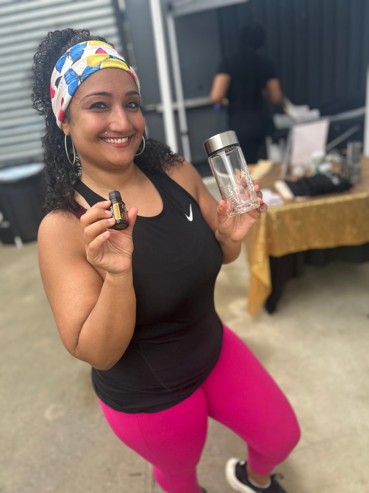 Smiling woman holding a small object and a glass, standing outdoors near a table with a gold tablecloth.