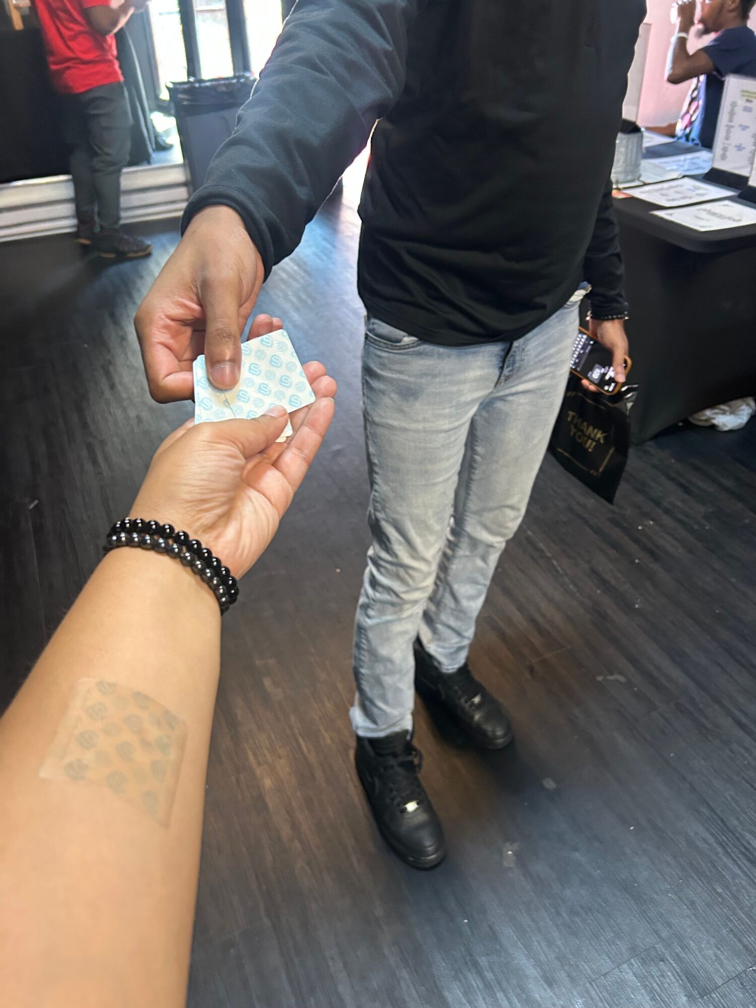 Person with a beaded bracelet hands a small card to another person with a bandage on their wrist, in a room with dark flooring.