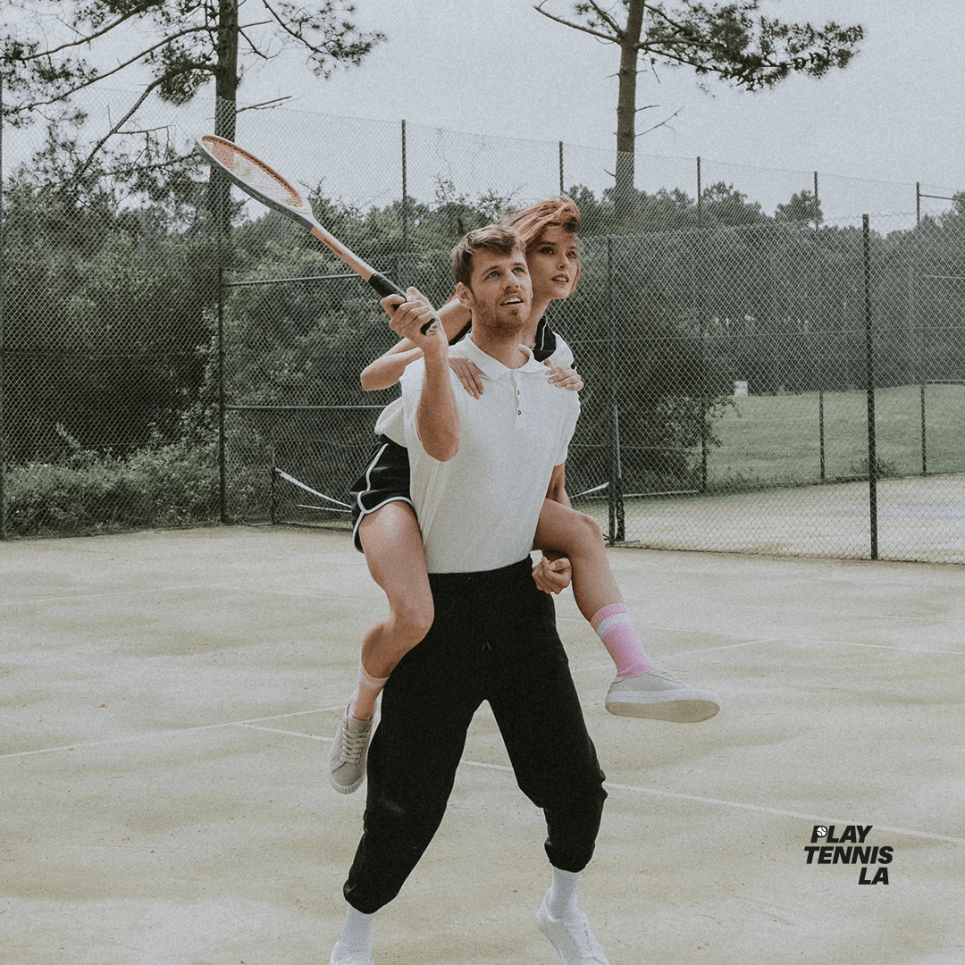 Person giving a piggyback ride to a child holding a tennis racket on a tennis court, trees in background.
