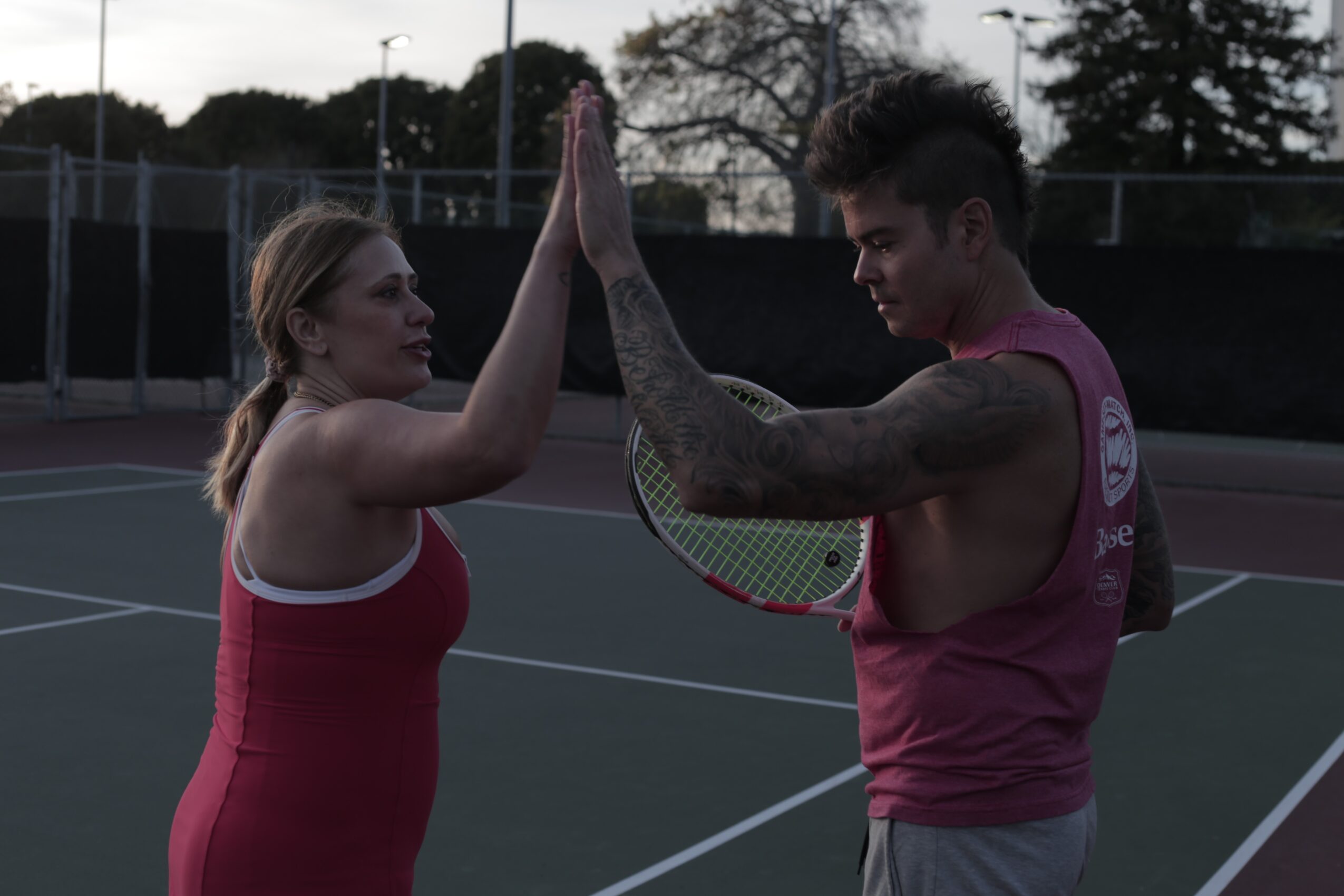 Two people on a tennis court giving high five, with trees and a fence in the background.