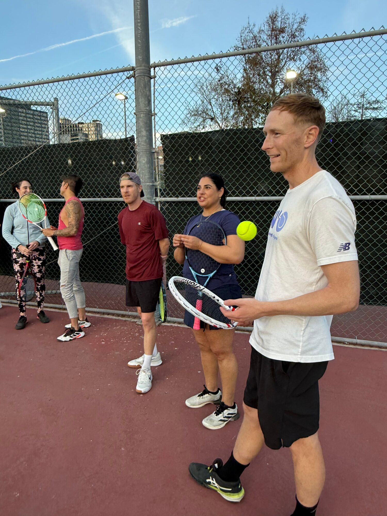 Group of five people on a tennis court, with a man holding a tennis racket and a yellow tennis ball, smiling.