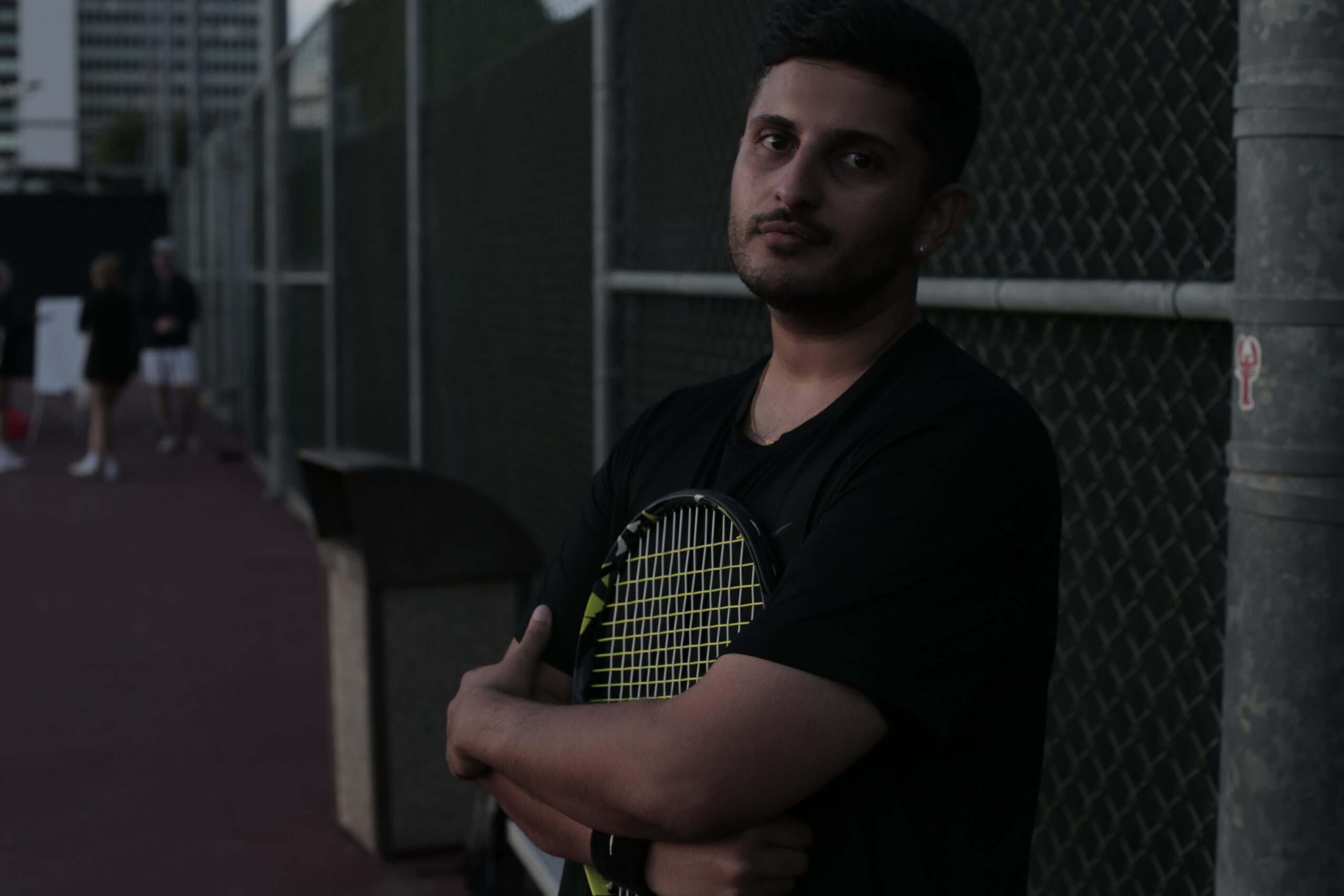Young man standing outdoors near a chain-link fence, holding a tennis racket, with a dark background and some people in the distance.