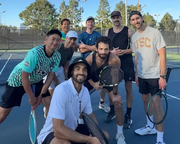 Group of nine people on tennis courts holding rackets, smiling, with trees and a fence in background.
