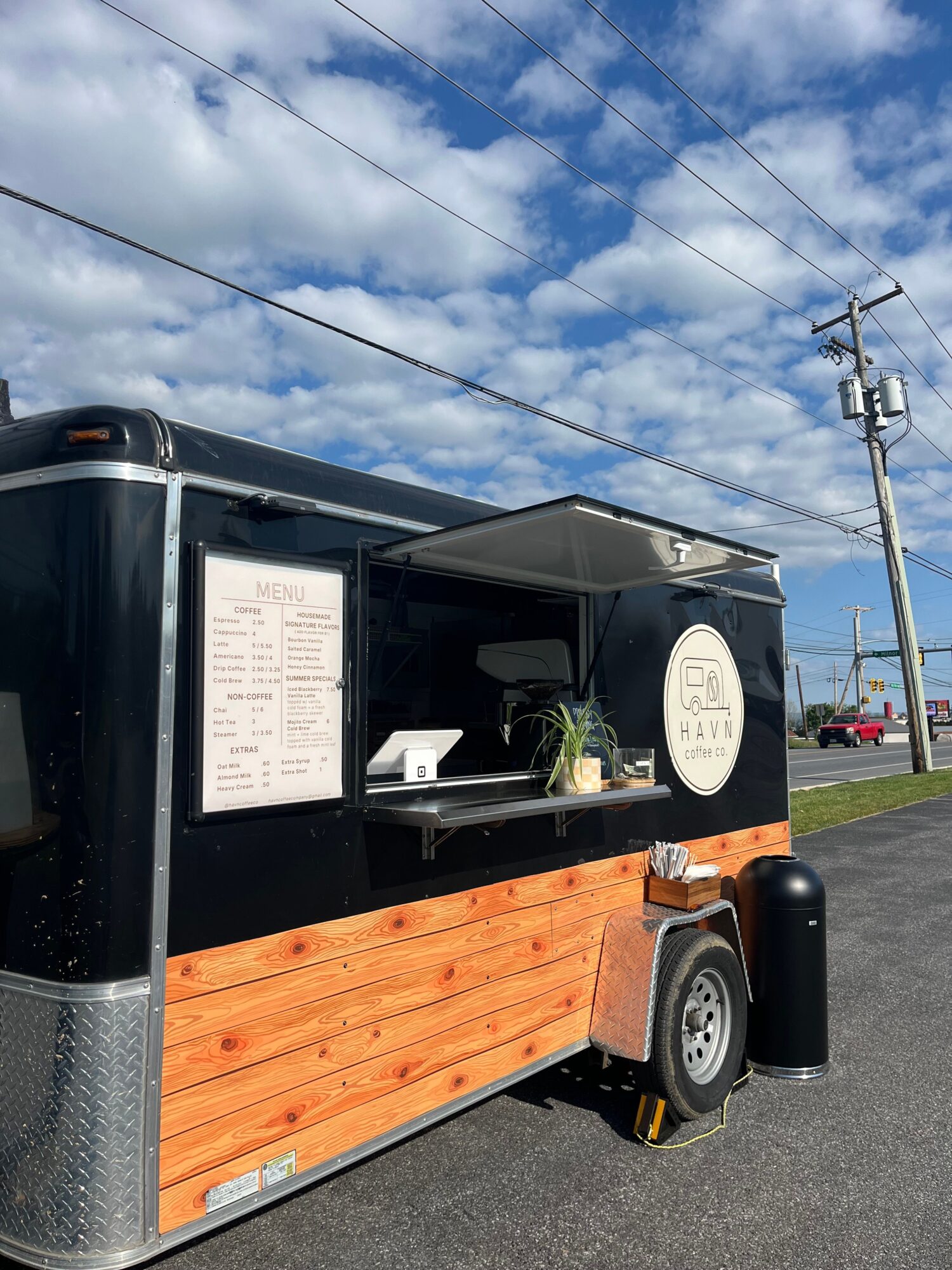 Food truck with black exterior, wooden serving area, menu, and a black trash can on a street with power lines and blue sky.