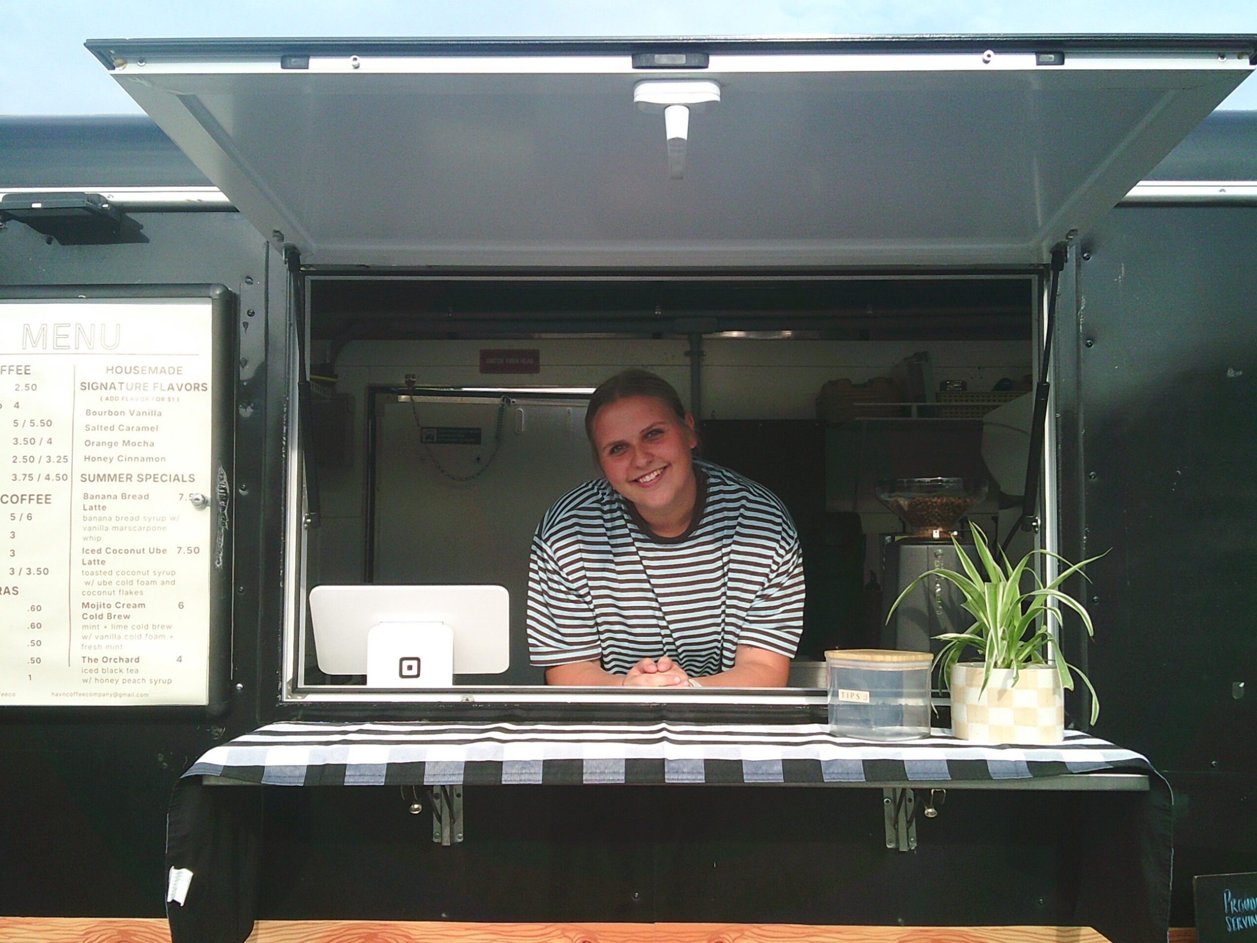 Person smiling behind a counter with a checkered tablecloth, a plant, and a cash register inside a food truck.
