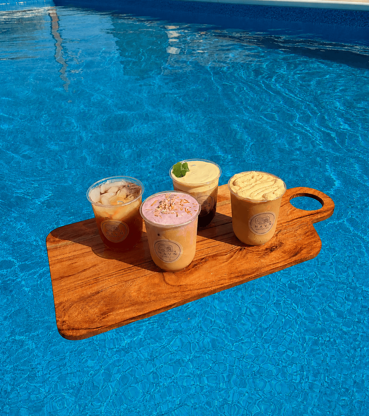 Four cups of ice cream on a wooden tray floating on a swimming pool, with blue water in the background.