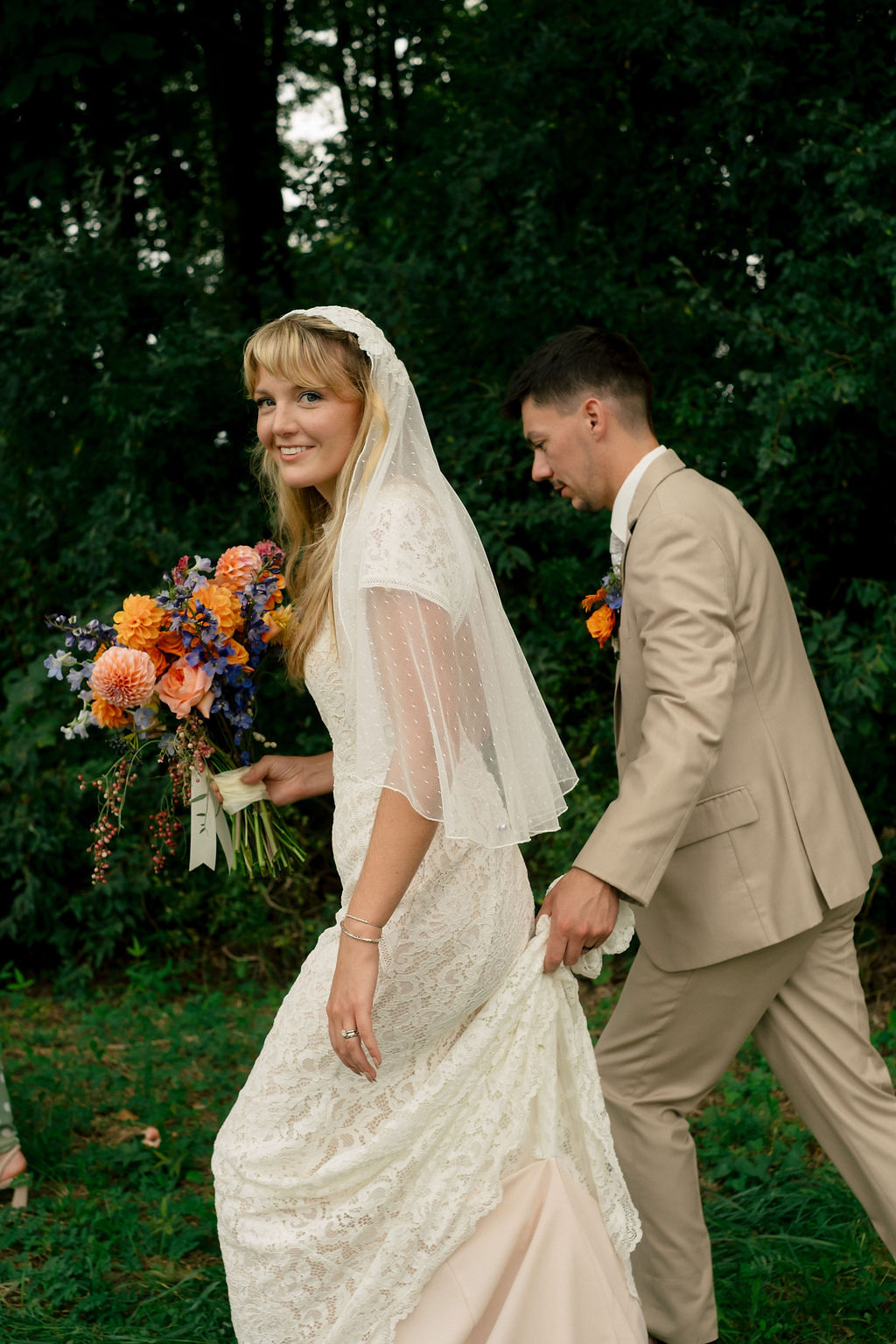 Bride in a white dress holding a bouquet, smiling, with groom in a beige suit walking behind her outdoors.