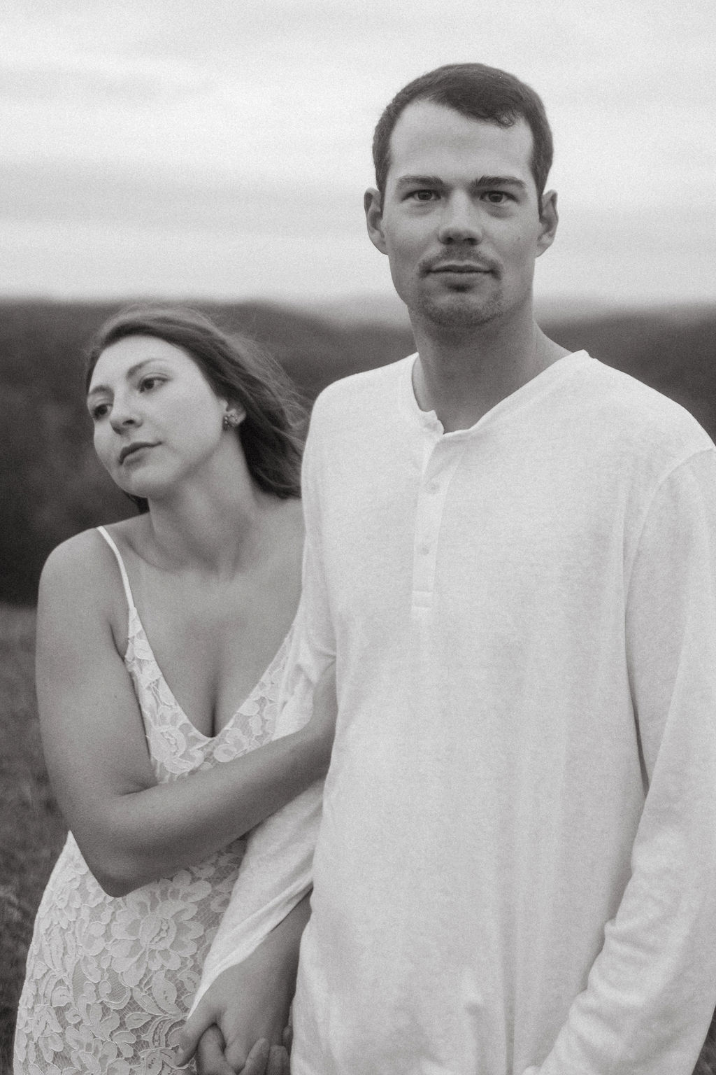 A man and woman stand outdoors, holding hands, with a landscape background. The woman looks at the man, who faces forward.