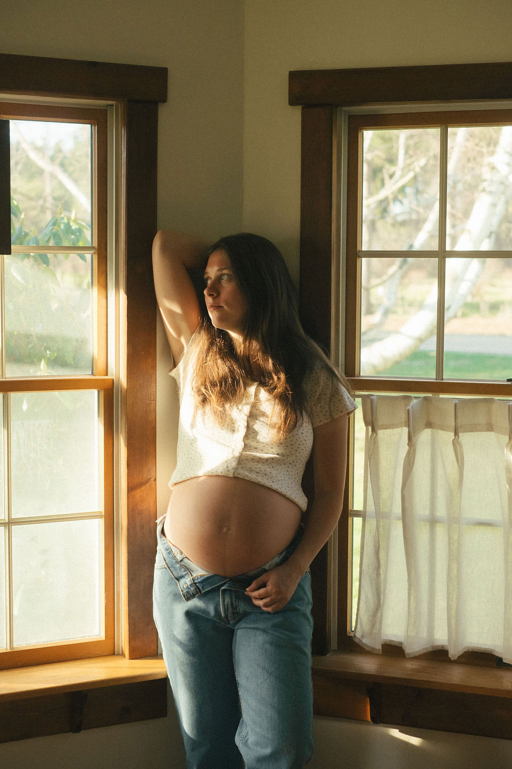 Pregnant woman standing by windows, sunlight on her face and belly, hand on her head, casual clothing, indoor setting.