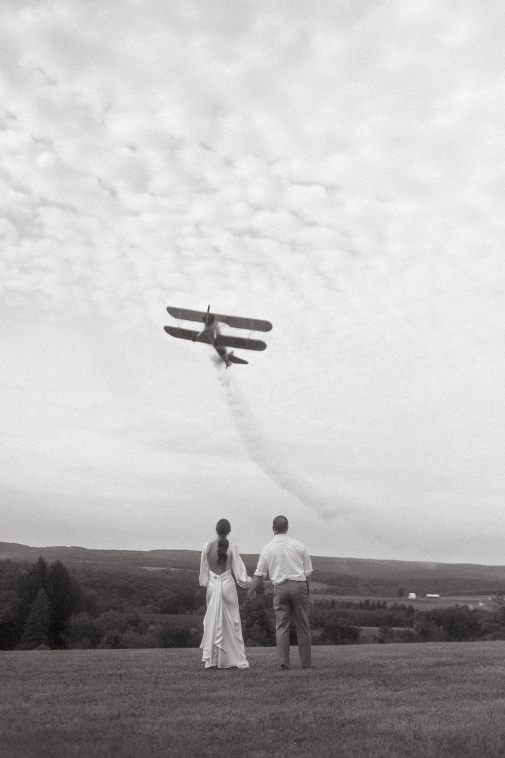 Two people stand on a hill watching a biplane fly overhead, leaving a trail of smoke in the sky, with clouds above.