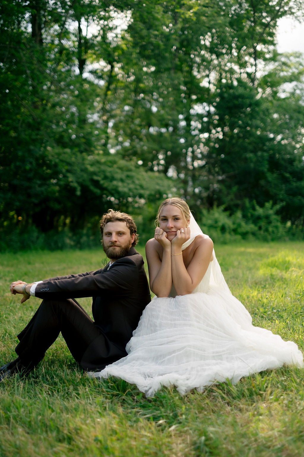 A couple sitting on grass in a park, with trees in the background, the woman in a white dress and the man in a dark suit.