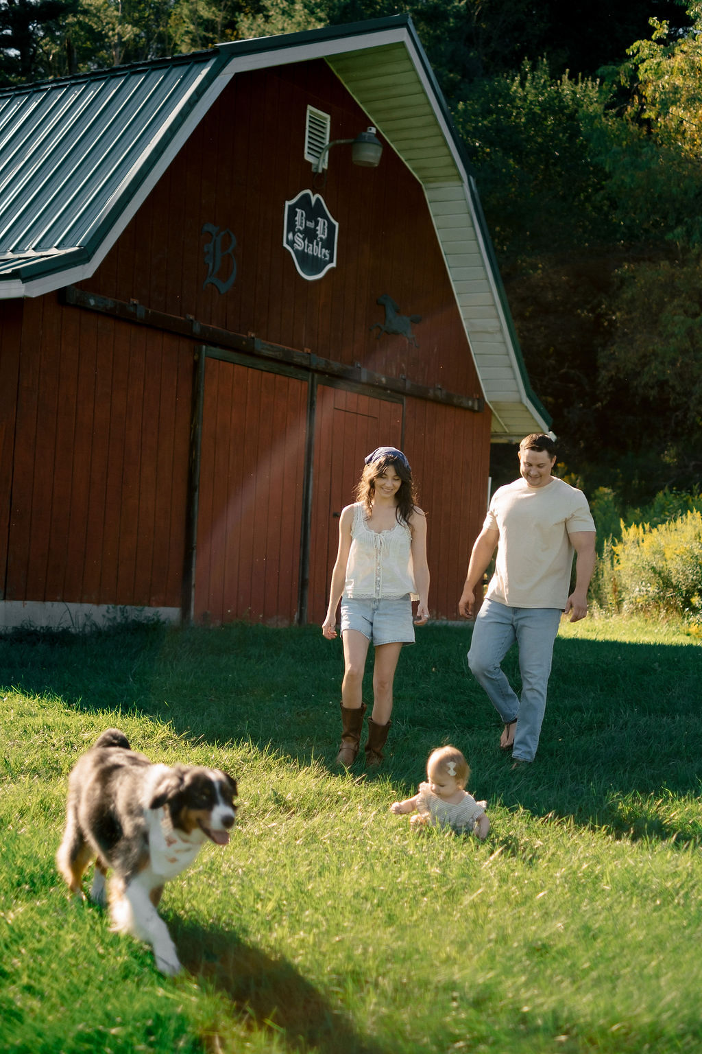 A couple walking on grass near a red barn with a sign, a dog running, and a baby sitting on the grass.