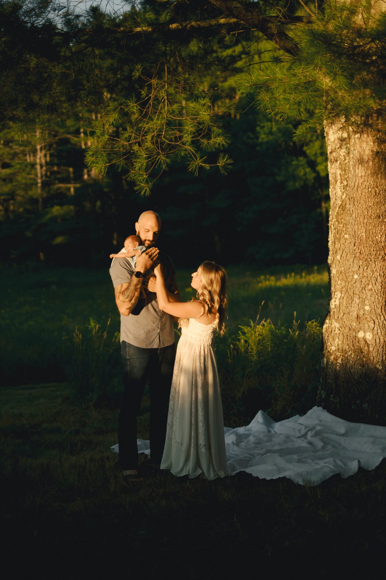A man and woman holding hands outdoors at sunset, with trees and grass in the background, and a white cloth on the ground.