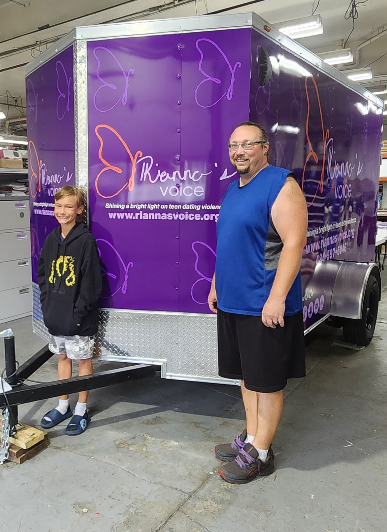 Two people standing in front of a purple mobile unit with text and graphics, indoors. One person is a child, the other an adult male.
