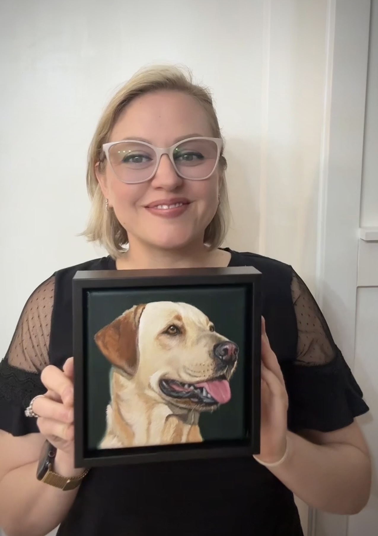 Woman with glasses holding a framed picture of a dog, smiling, indoors.