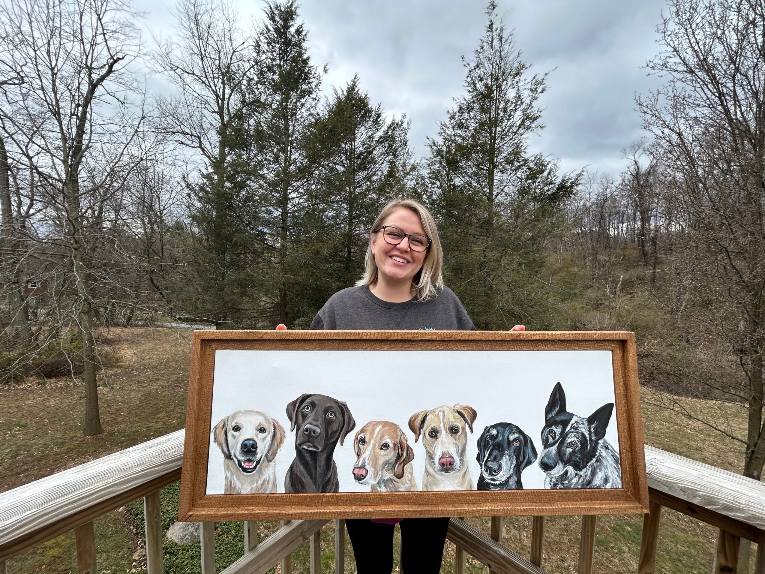Woman smiling outdoors holding a framed painting of seven dogs, with trees and cloudy sky in background.