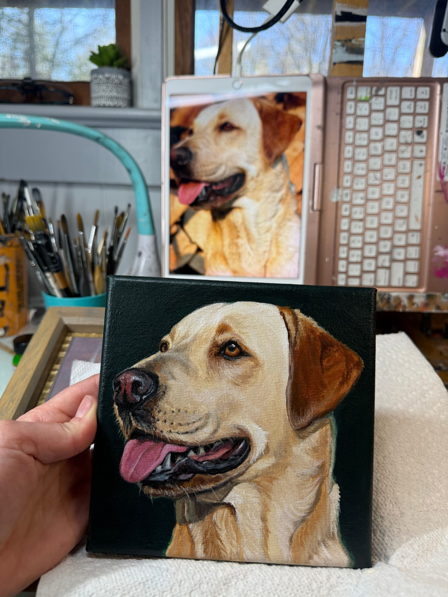 Hand holding a small canvas painting of a dog with its tongue out, with a computer and a photo of the same dog in the background.