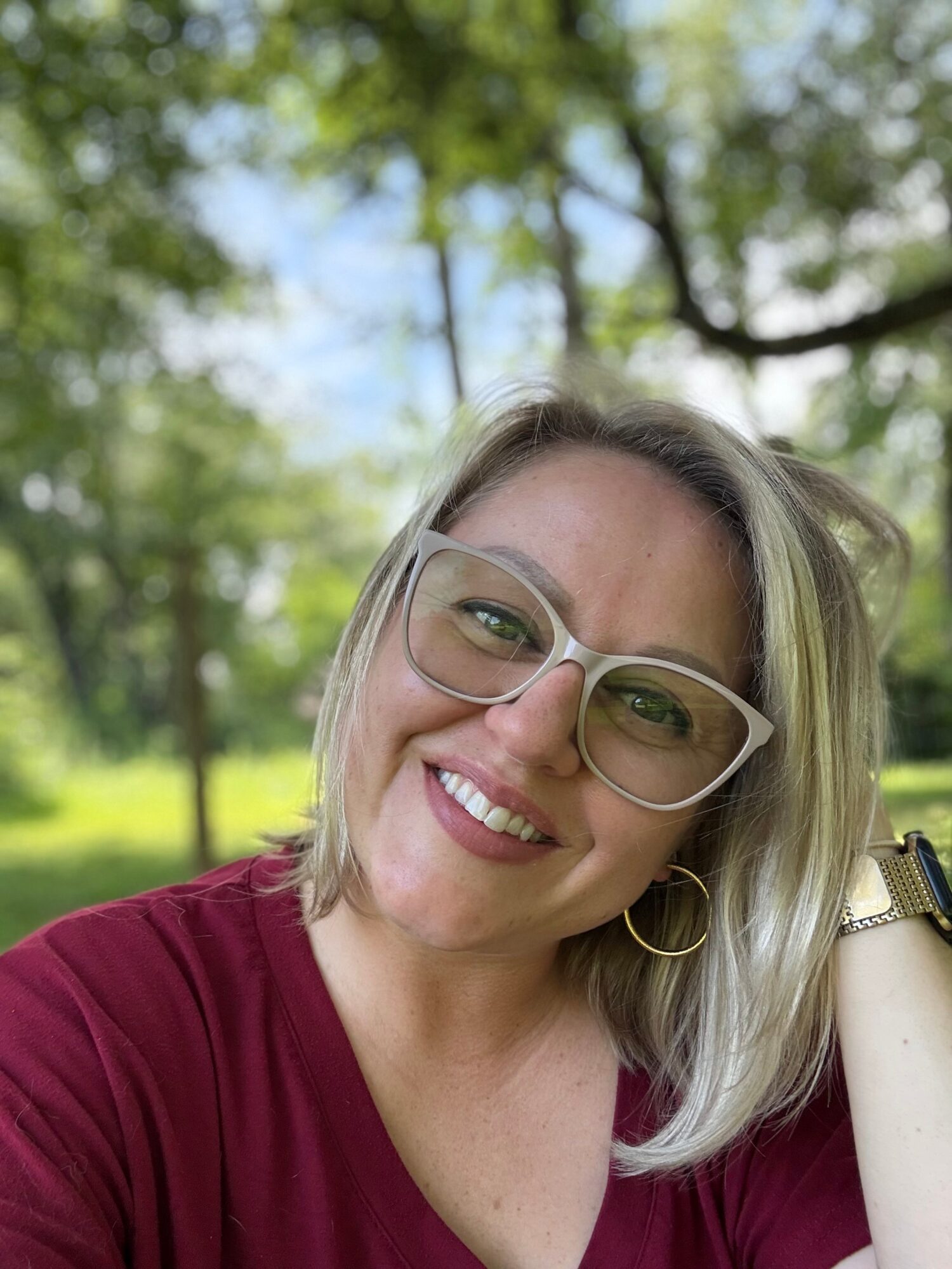 Woman with glasses smiling outdoors, trees and blue sky in background.