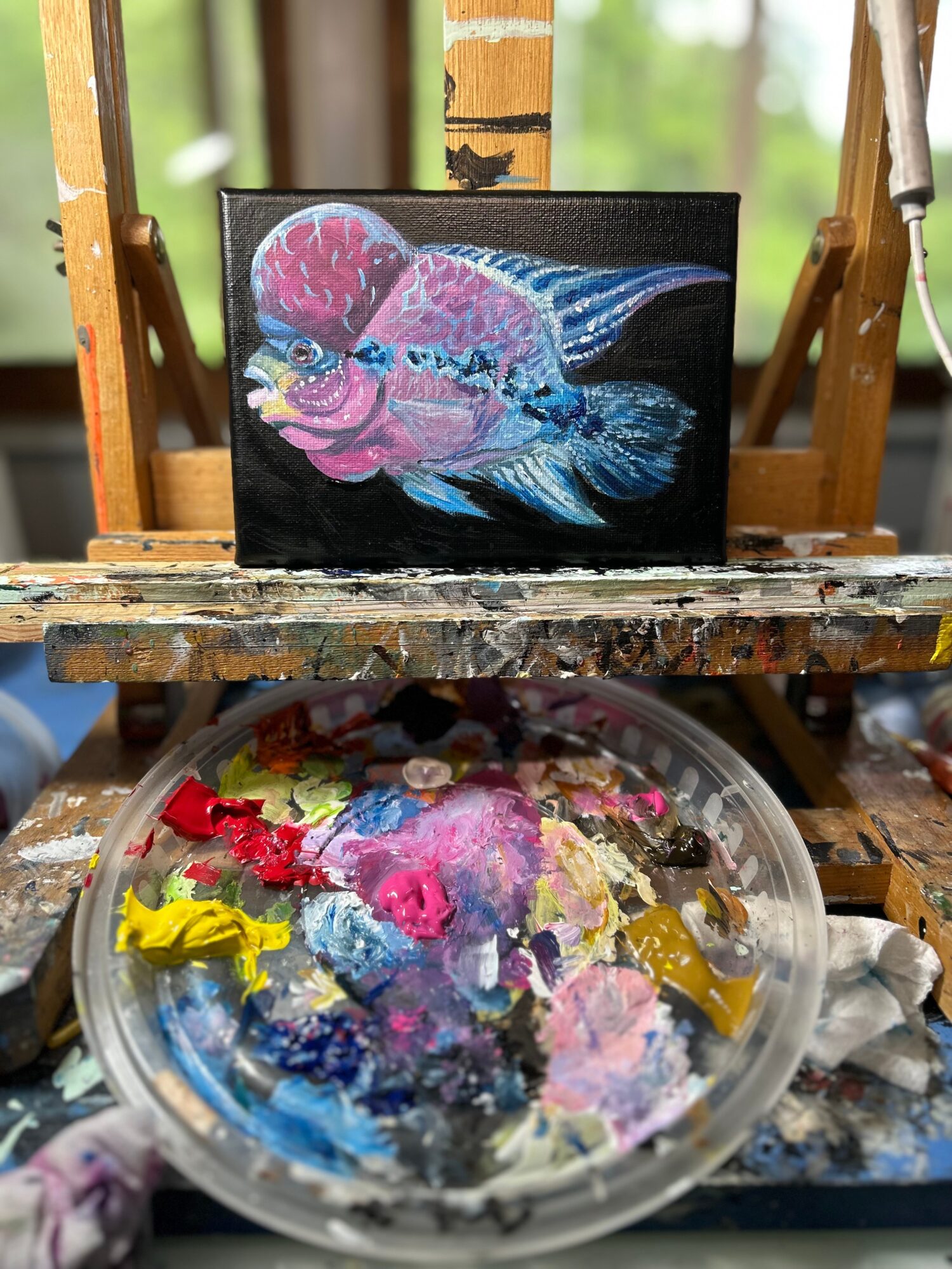 Painted fish artwork on a black canvas, placed on an easel, with a palette of mixed paints below, in a bright room.