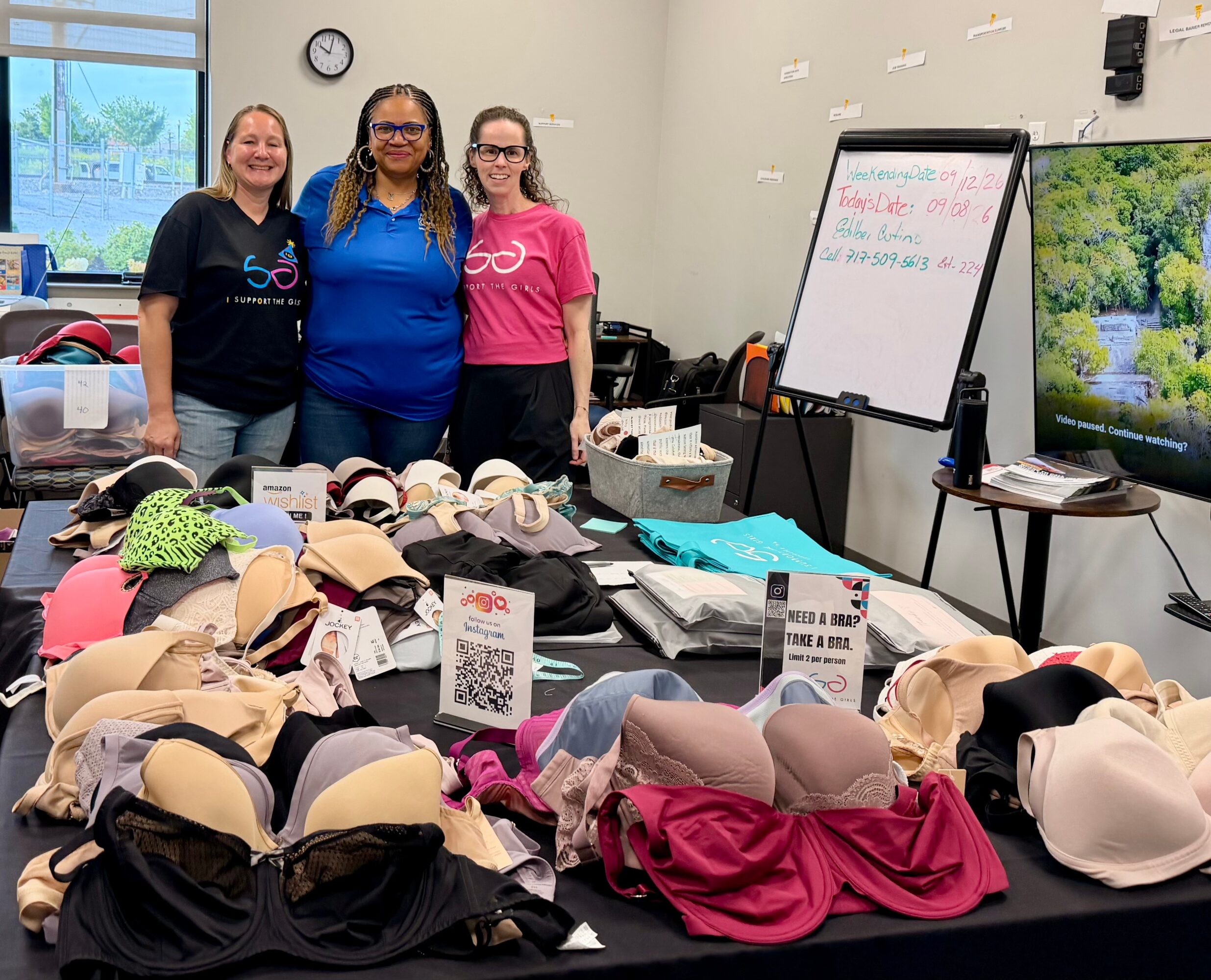 Three women stand behind a table filled with bras and hats in a room with a whiteboard and a window.
