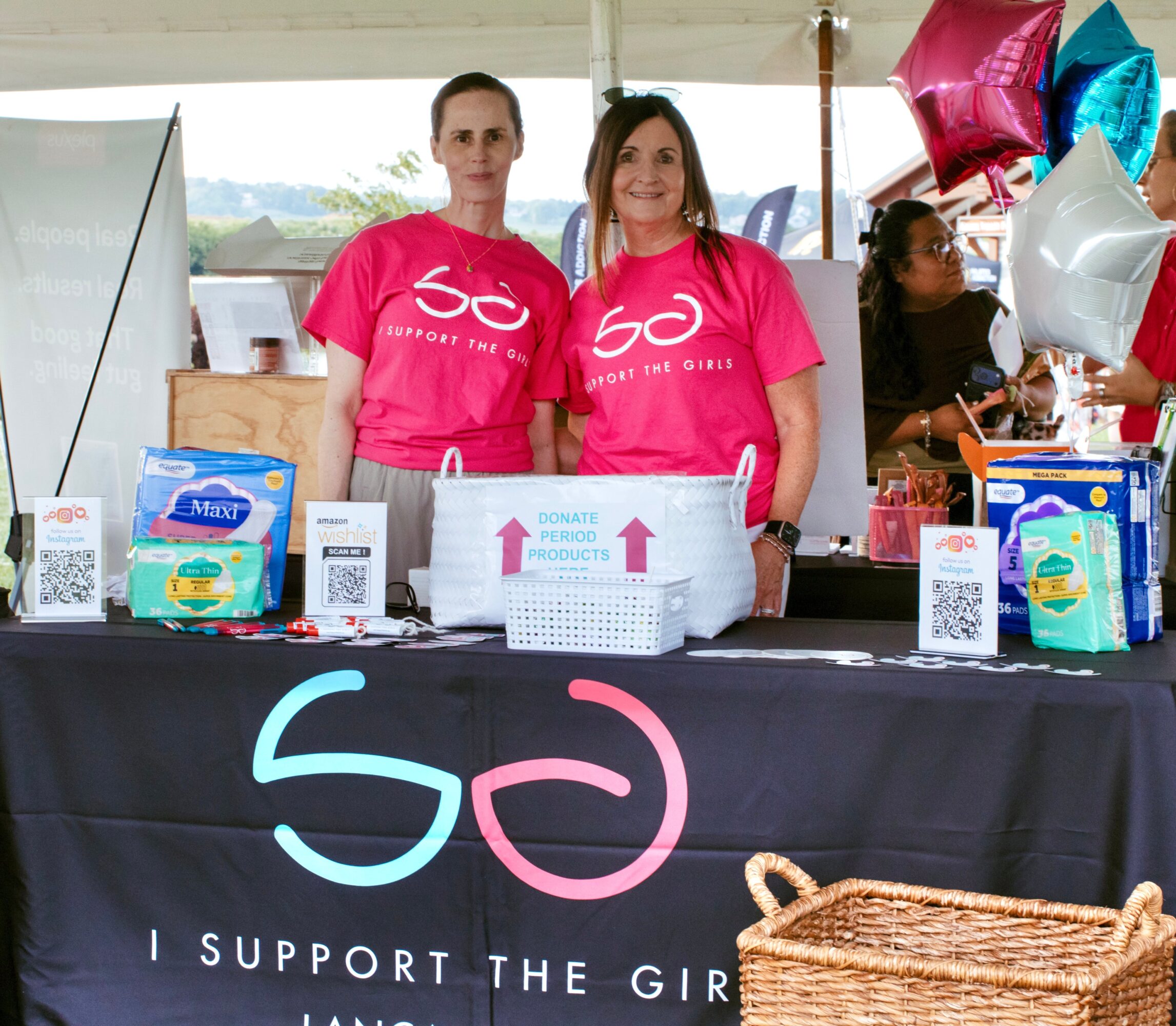 Two women in pink shirts stand behind a table with support products and balloons, at an outdoor event.