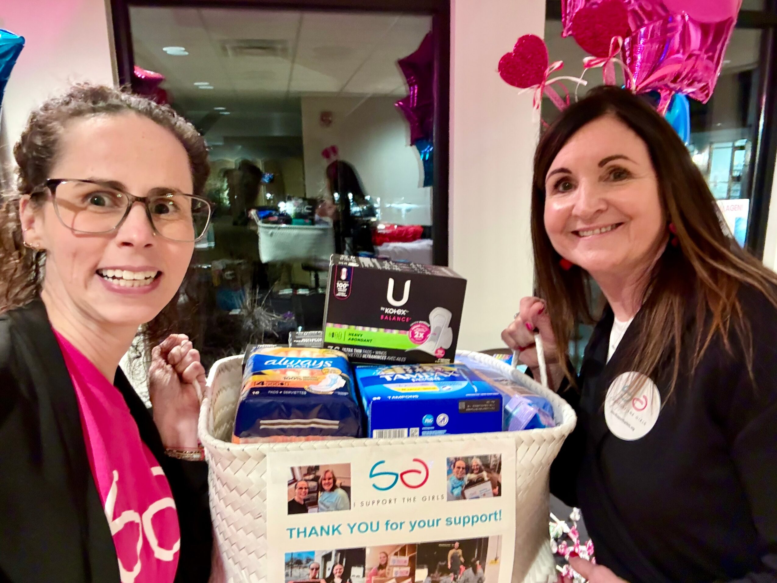 Two women smiling behind a gift basket with a thank you note, celebrating a 50th support anniversary, decorated with pink hearts.