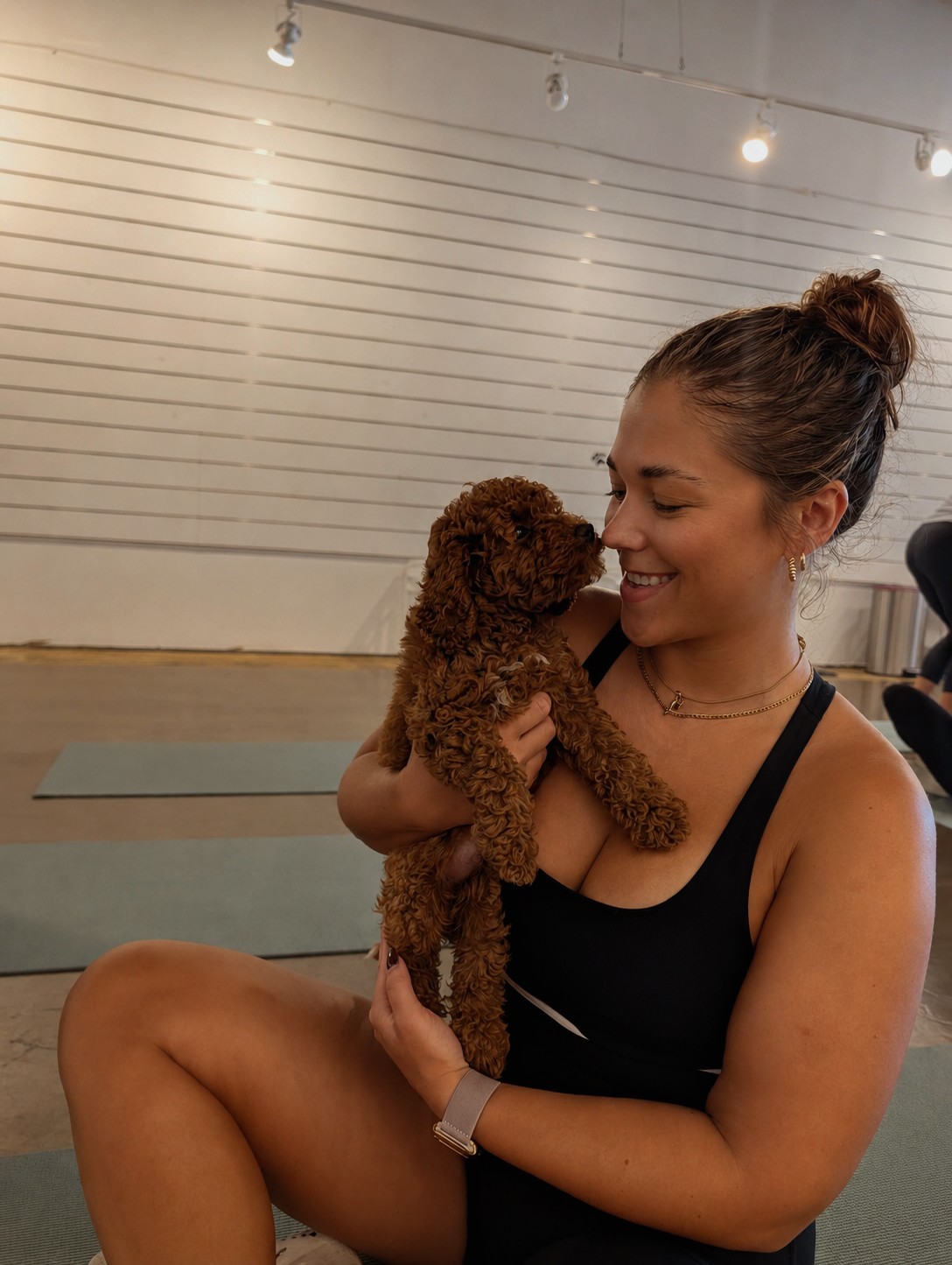 Woman holding a small curly-haired dog, smiling, indoors with white wall and string lights in background.