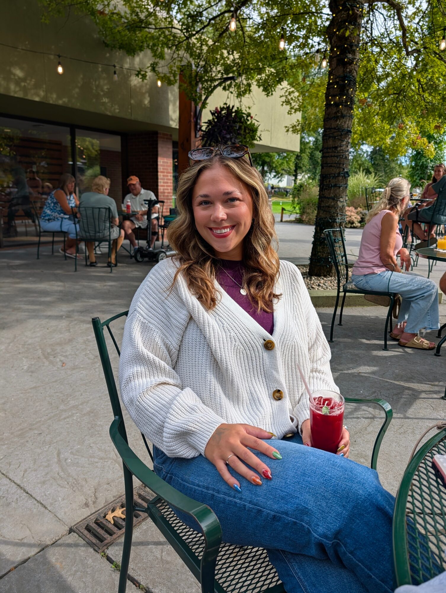 Woman sitting outdoors at a cafe, smiling, holding a drink, with trees and other people in the background.