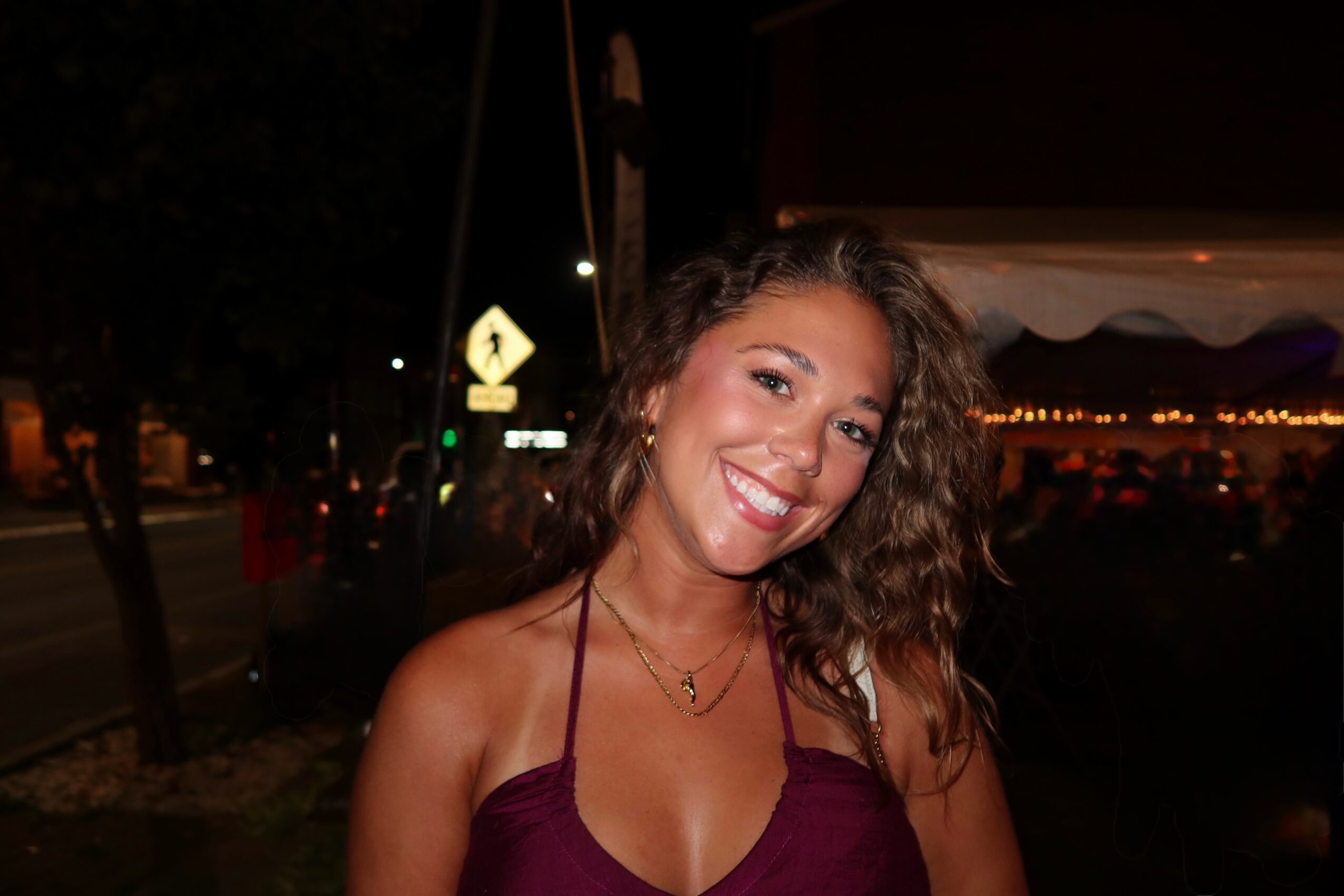 Young woman with curly hair smiling outdoors at night, wearing a maroon top, in a lively setting.