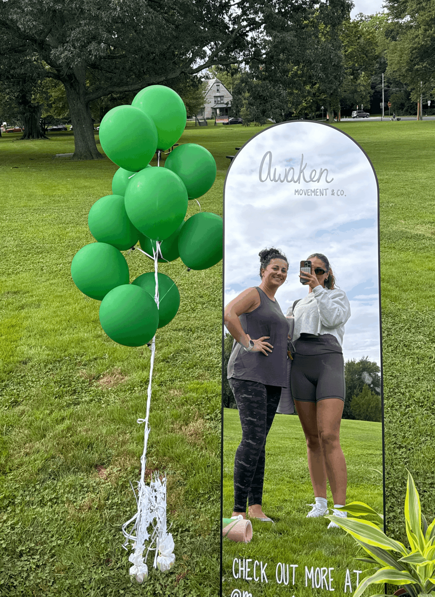 Two women standing outdoors near a reflective sign with balloons attached, trees and a house in the background, sunny day.