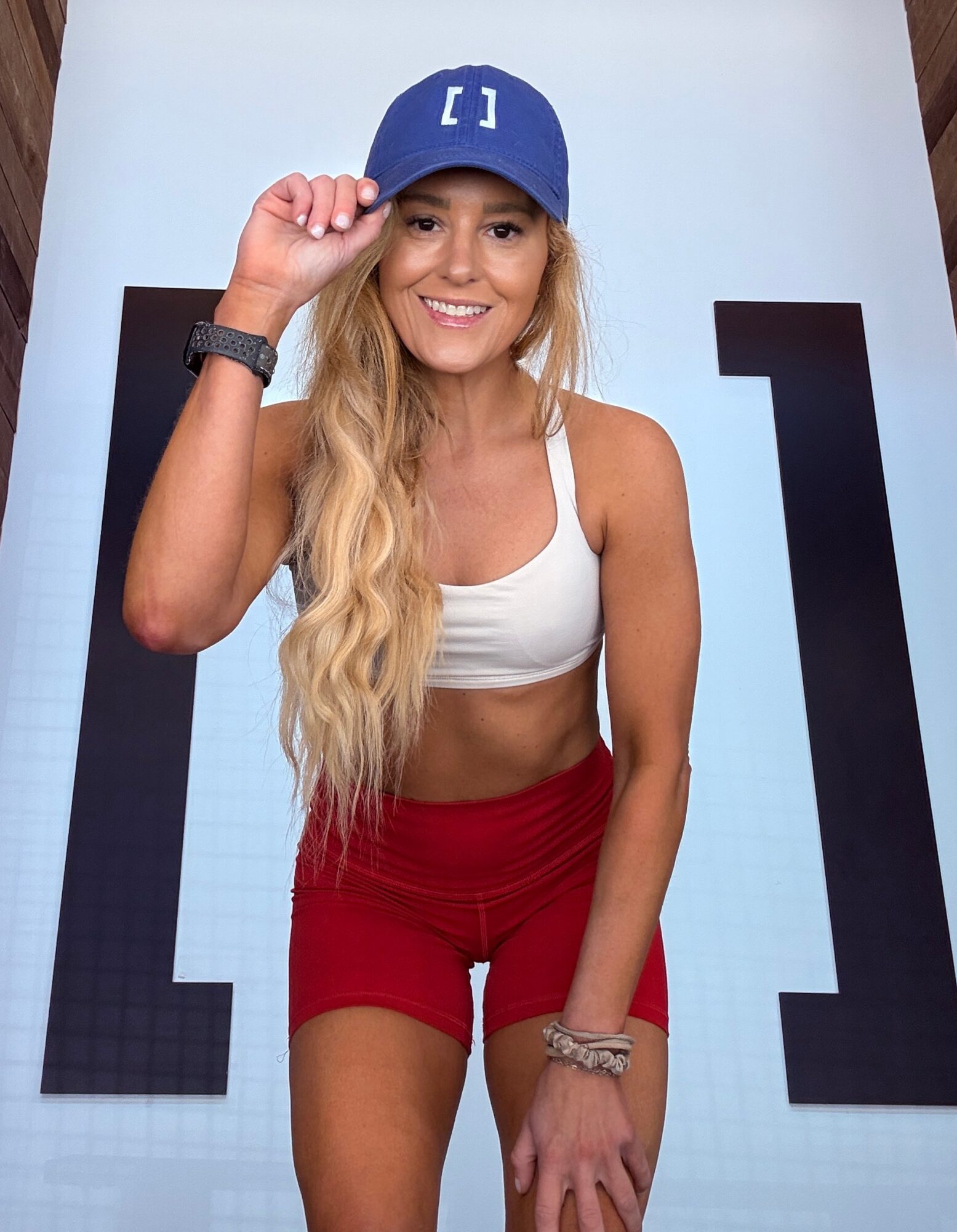 Young woman with long blonde hair wearing a blue cap, white sports bra, and red shorts, smiling and holding her cap.