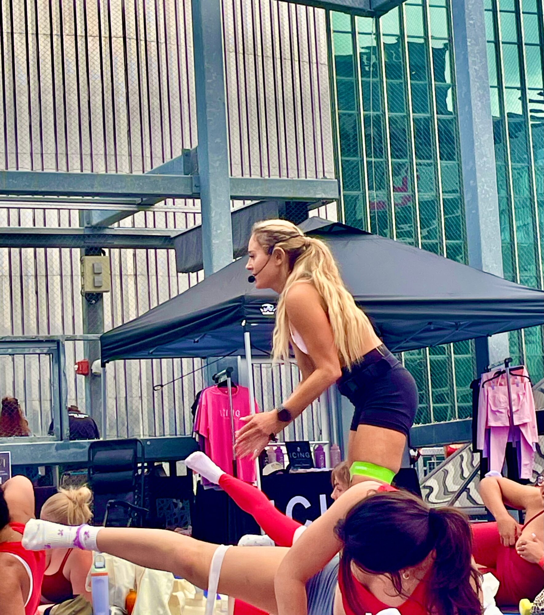 Young woman with long blonde hair in a black top and shorts, performing a dance move outdoors with children nearby.