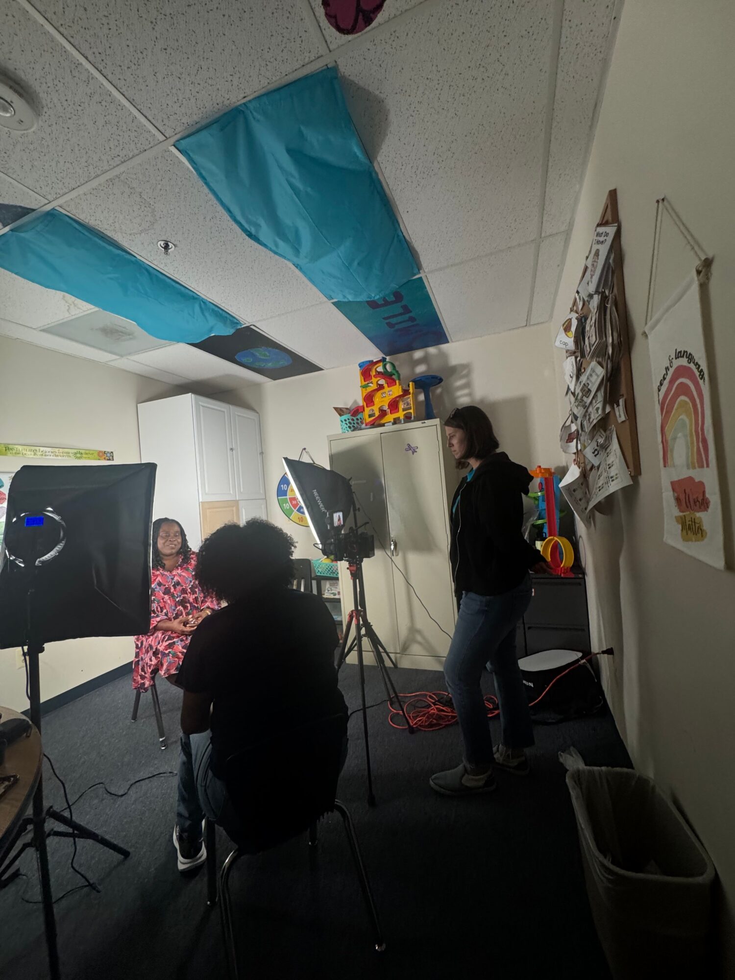 People in a room with a white wall, colorful artwork, and a person using a computer. Two people are standing and one is sitting.