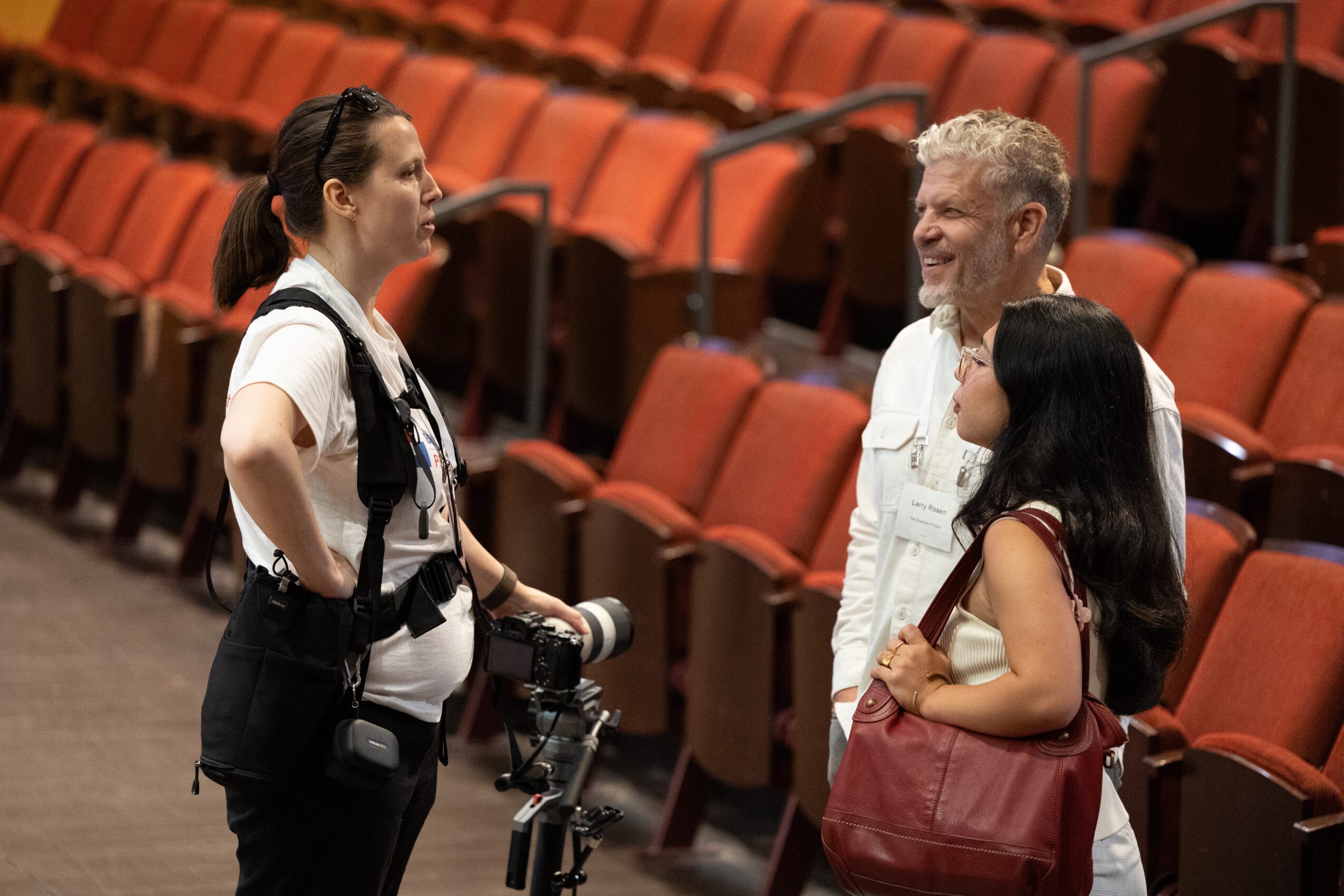 Three people standing and talking in an auditorium with orange seats, two women and one man smiling and engaging.