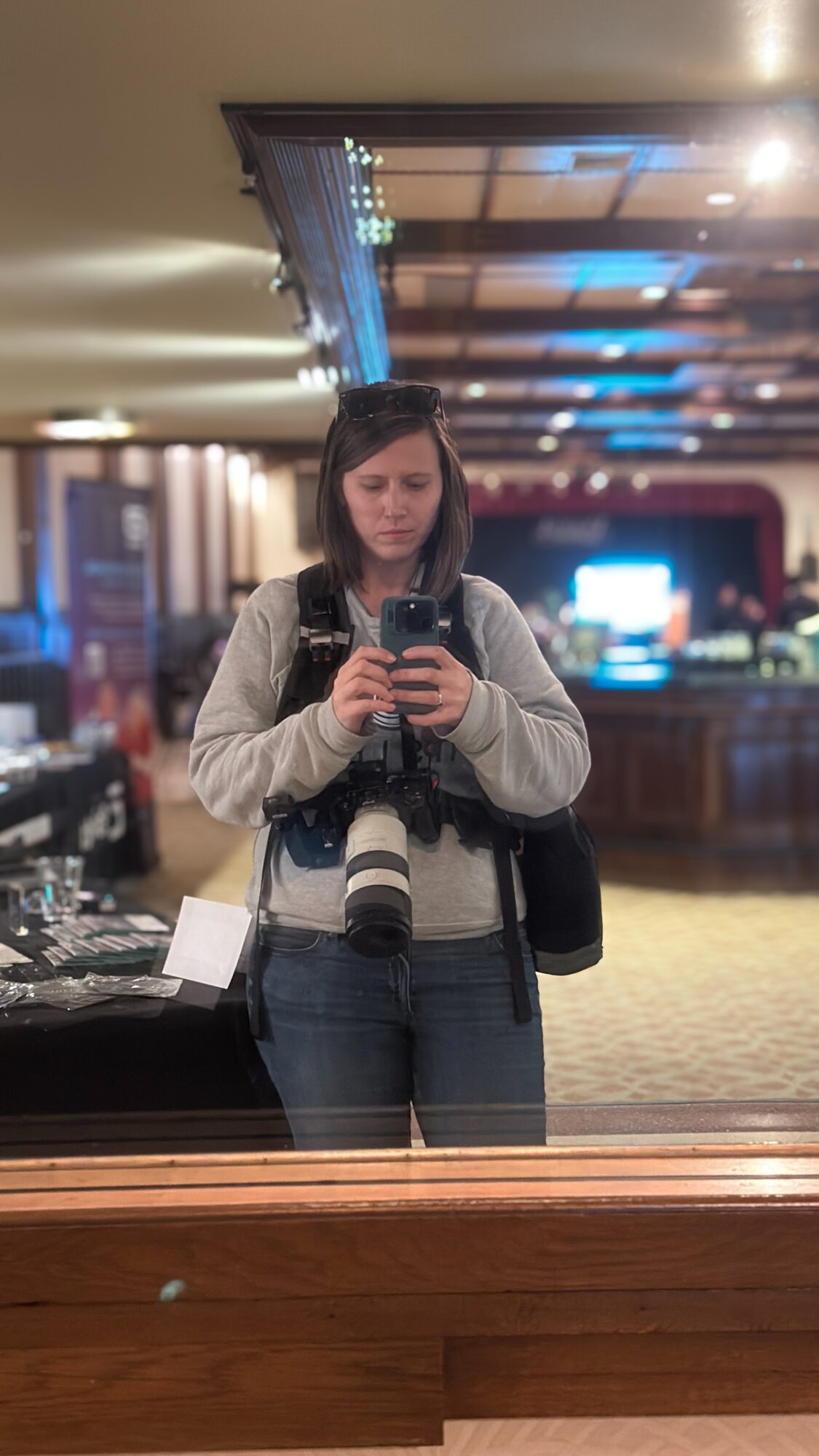 Young woman taking a mirror selfie in a hotel lobby, holding a camera with a large lens, wearing a gray jacket and jeans.