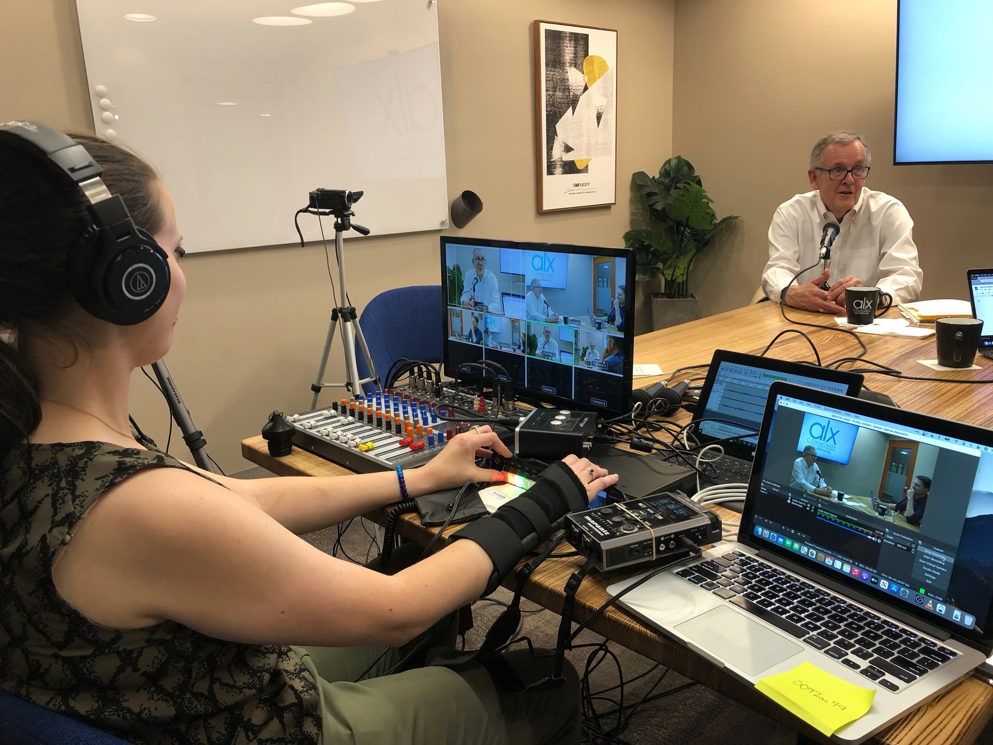 Woman with headphones operating video equipment in a conference room with a man speaking at the table.